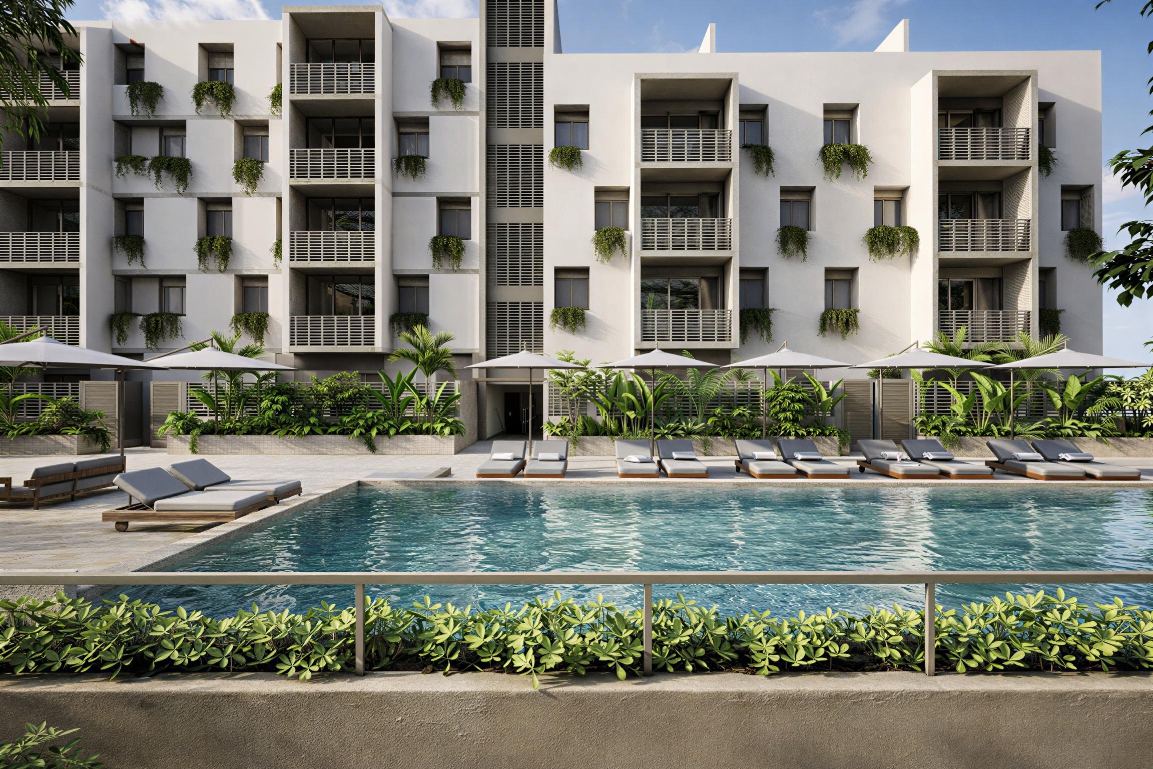 Vista de un edificio moderno con varias plantas, balcón y ventanas, rodeado de vegetación y plantas decorativas, con piscina y tumbonas en primer plano.
