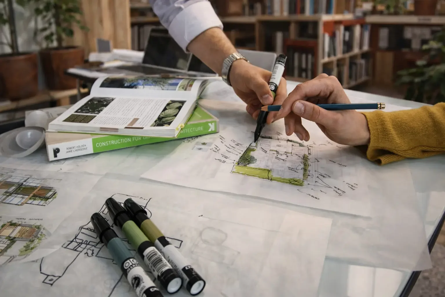 Dos personas trabajan en planos y bocetos arquitectónicos sobre una mesa, con marcadores y una revista de arquitectura a un lado.