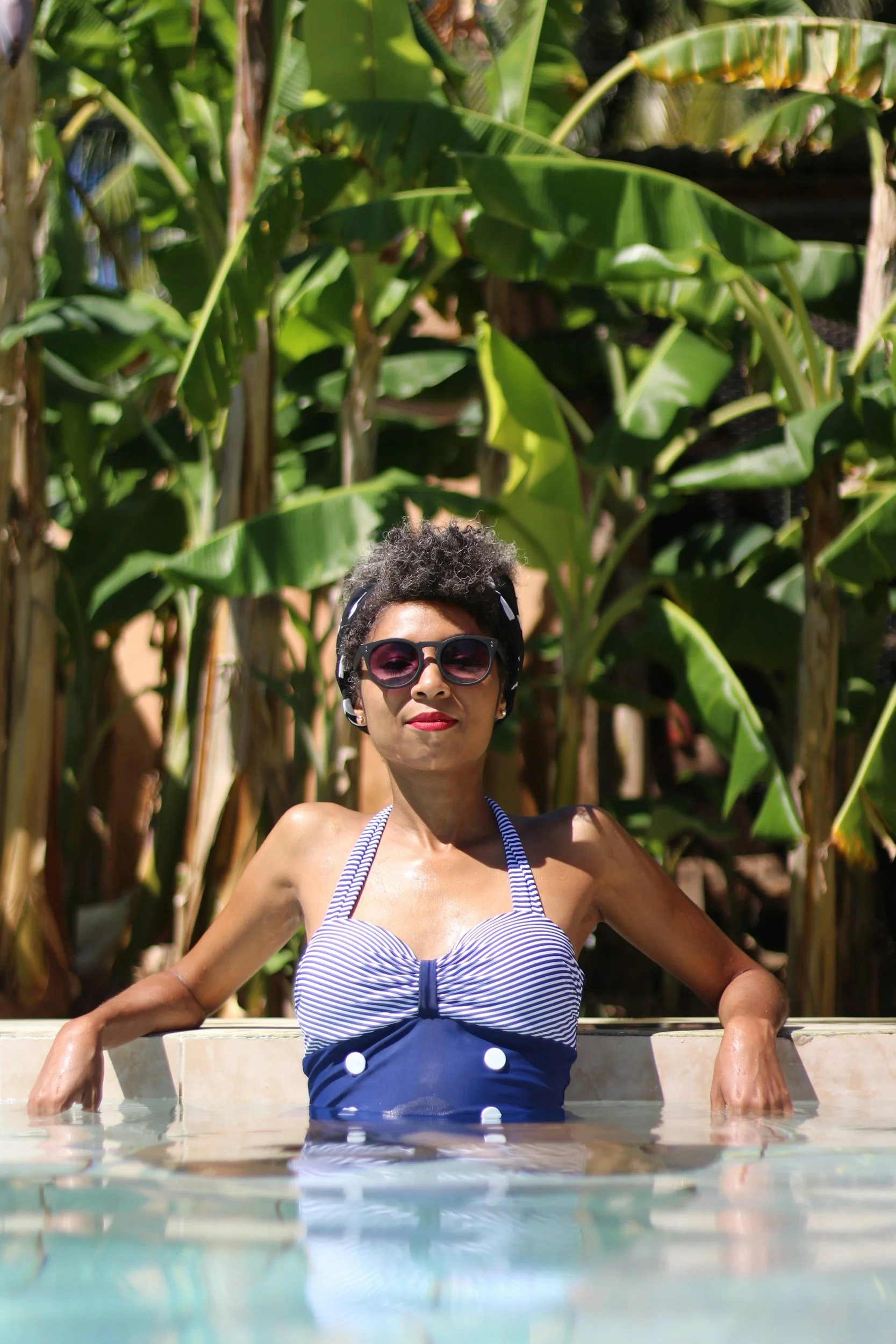 Woman with sunglasses relaxing in a pool with lush green banana leaves in the background.