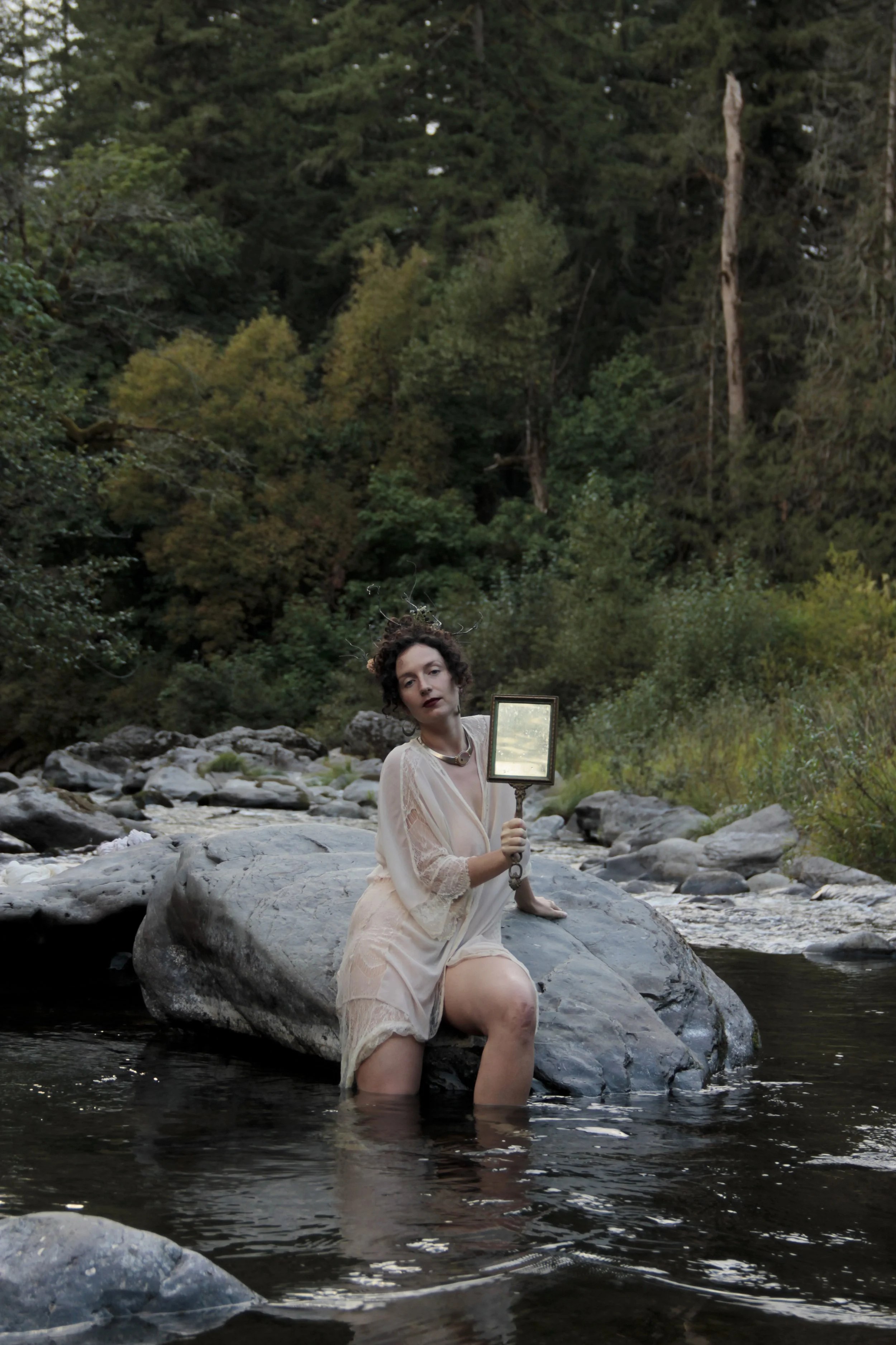 A woman in a soft, light dress sitting on a large rock in a shallow stream, holding a mirror in her right hand, surrounded by dense green forest.
