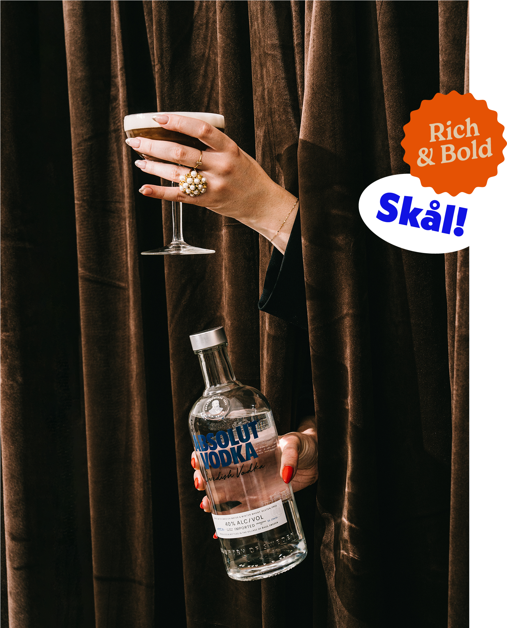 Hand holding a martini glass with a dark drink, jewelry including a large pearl ring, and a gold bracelet, partially visible behind brown velvet curtains, with a hand holding a bottle of Absolut vodka.