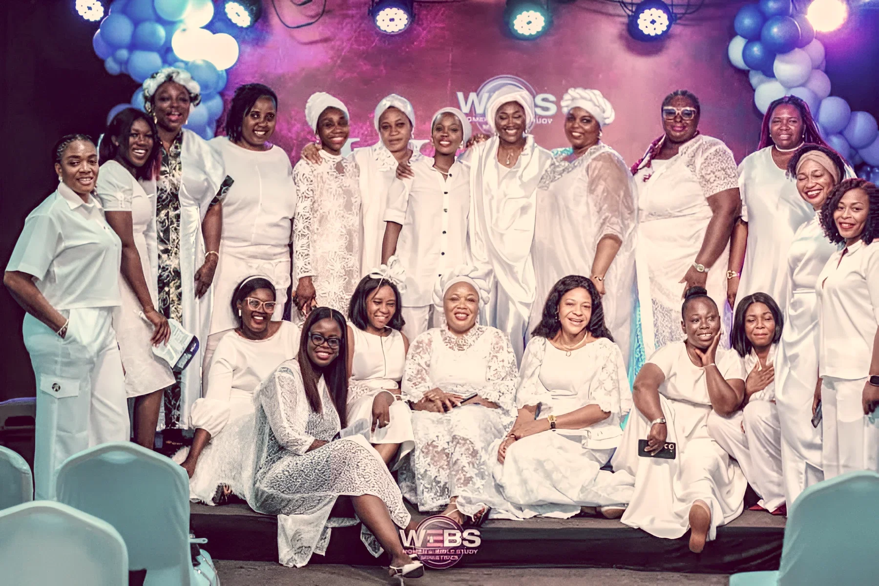 Two women in vibrant African dresses pose outdoors against a natural background; one wears a dark, sequined gown, and the other wears a bright red dress with gold patterns and a headscarf.
