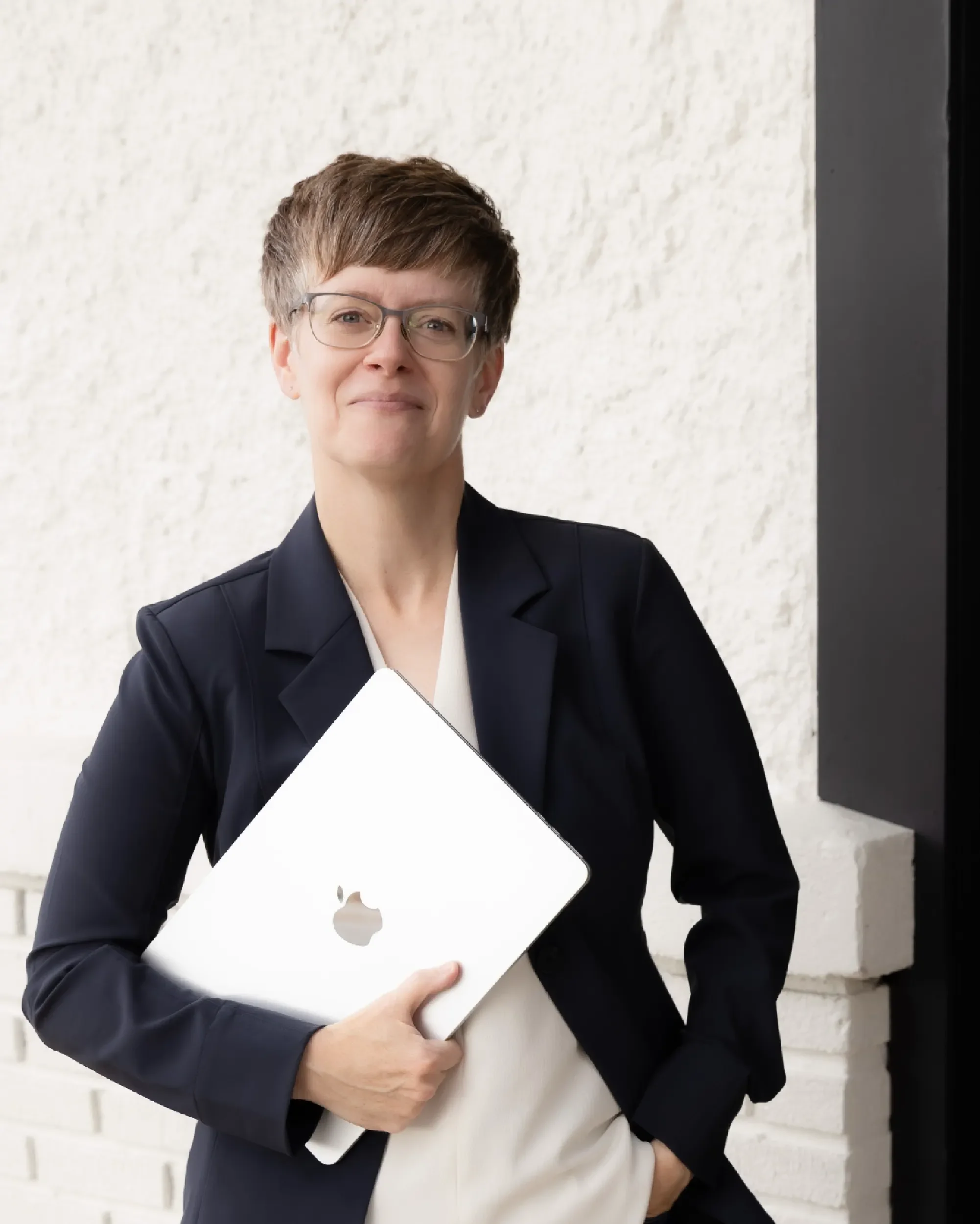 A professional woman wearing glasses and a dark blazer, holding a silver MacBook, standing outside against a beige textured wall.