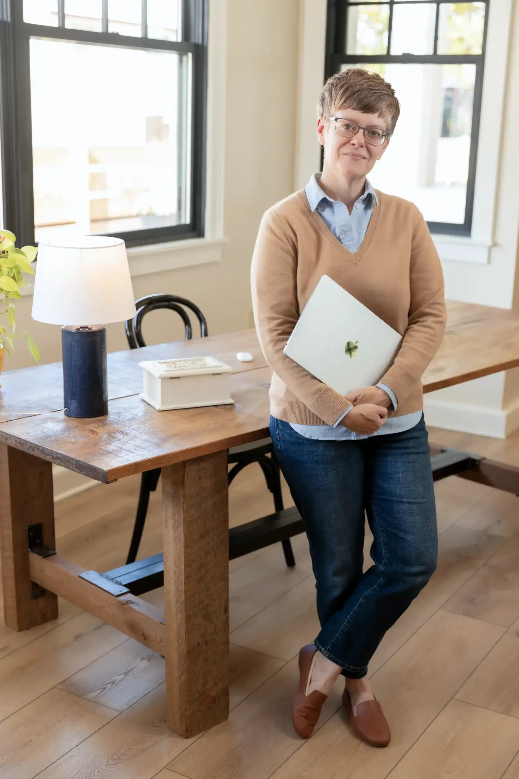 A woman wearing a beige sweater and a blue button down shirt leaning against a wooden desk and holding an Apple MacBook laptop