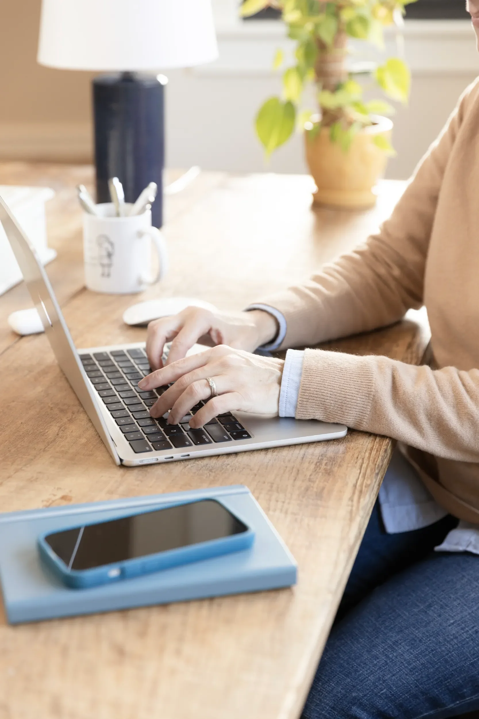 Person typing on a silver laptop with a smartphone in a blue case on top of a closed notebook on a wooden desk, with a white mug holding pens, a white mouse, a black water bottle, and a potted plant in the background.