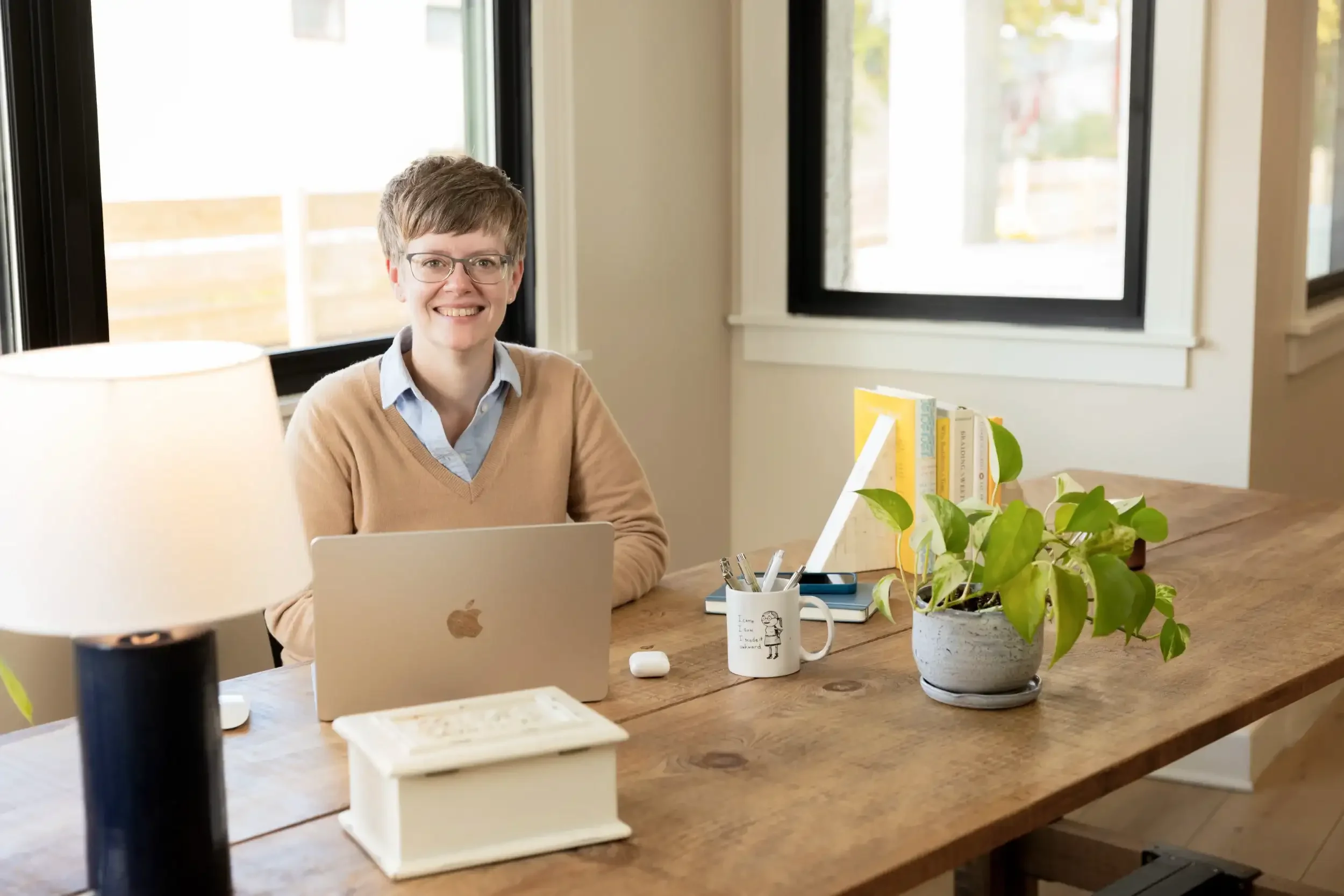 A smiling woman with short hair and glasses sitting at a wooden desk with a laptop, surrounded by office supplies, books, and a potted plant, in a bright room with windows behind her.