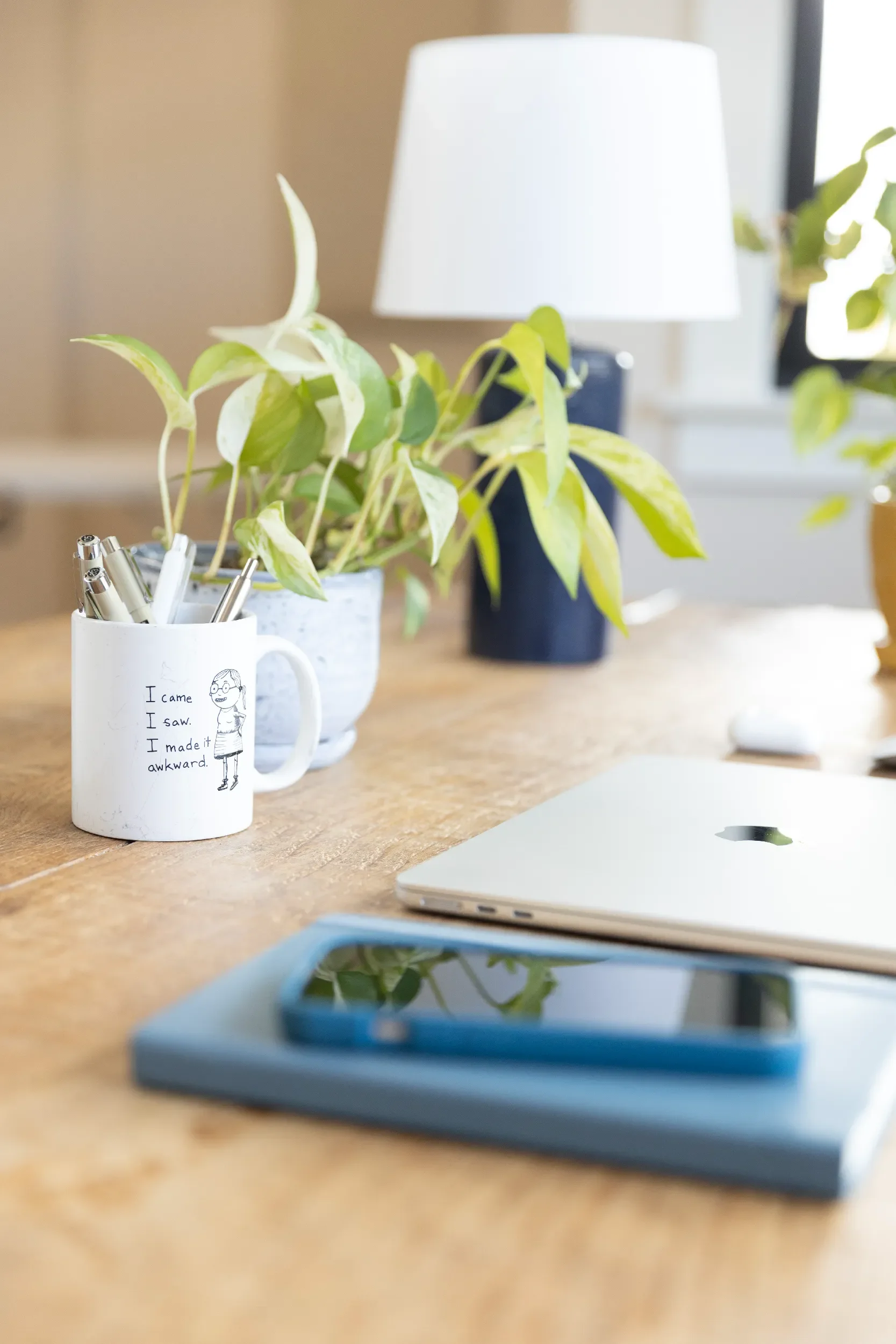 A tidy wooden desk with a white ceramic mug with a cartoon girl and handwriting, two potted plants, a closed silver laptop, and a smartphone in a blue case on a blue notebook.