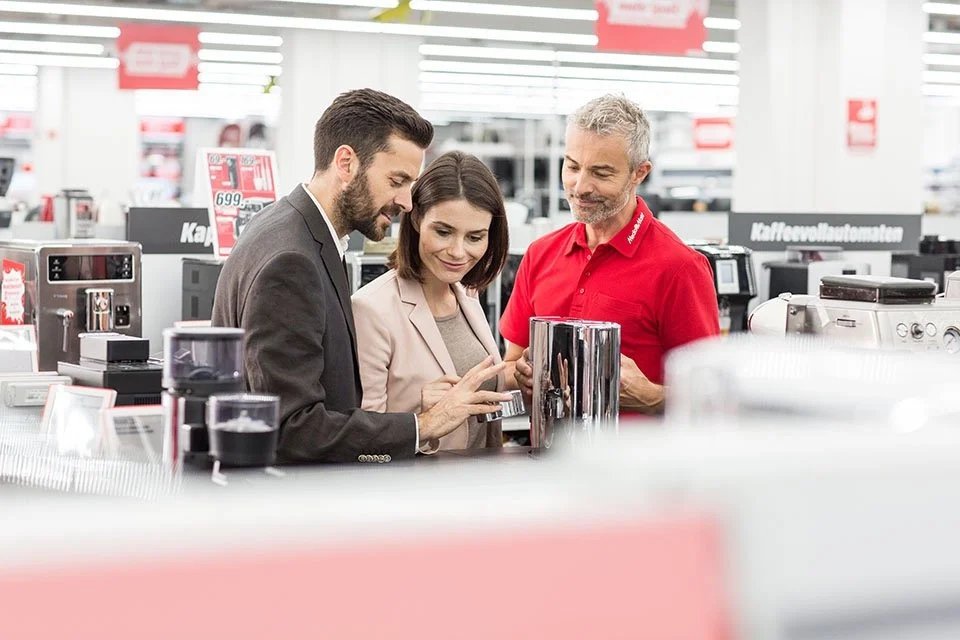 Drei Menschen, zwei Männer und eine Frau, stehen in einem Elektronikgeschäft vor einem Display mit Smartphones. Der Mann auf der linken zeigt auf sein Smartphone, während die anderen beiden zuschauen.