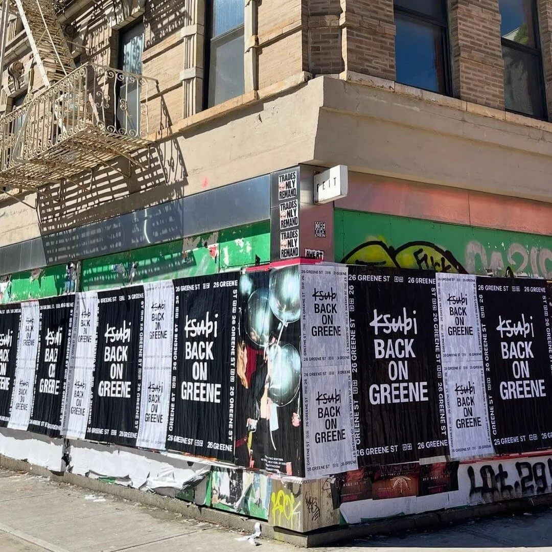 Street corner with posters reading 'Ksubi Back on Greene' attached to a wall, graffiti, a fire escape, and building windows above.