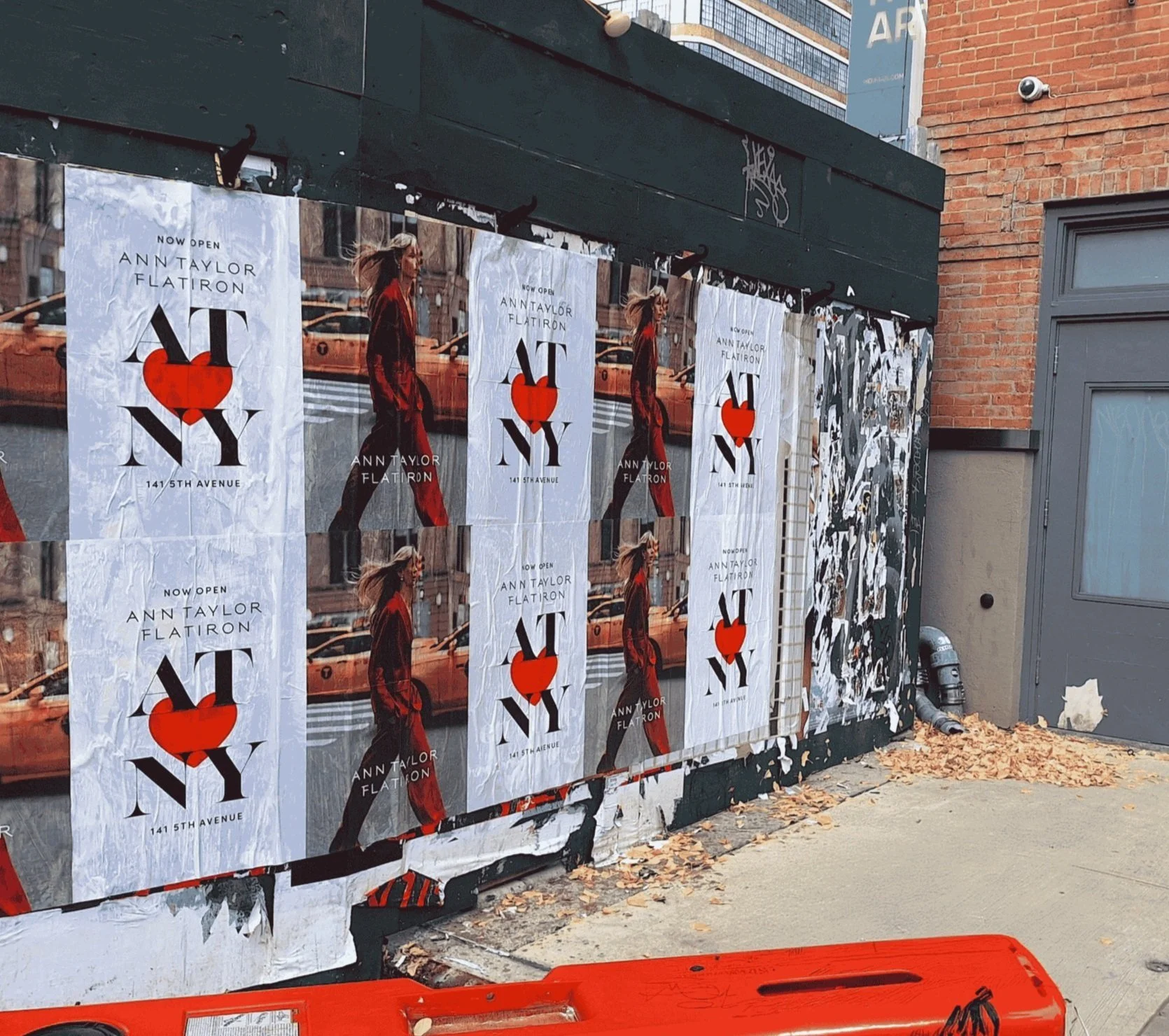 Street scene with posters advertising an event at 141 5th Avenue featuring Ann Taylor Flatiron, with a woman walking across a city street printed on the posters. The posters are peeling off a billboard on a sidewalk near a brick building. Fallen leav