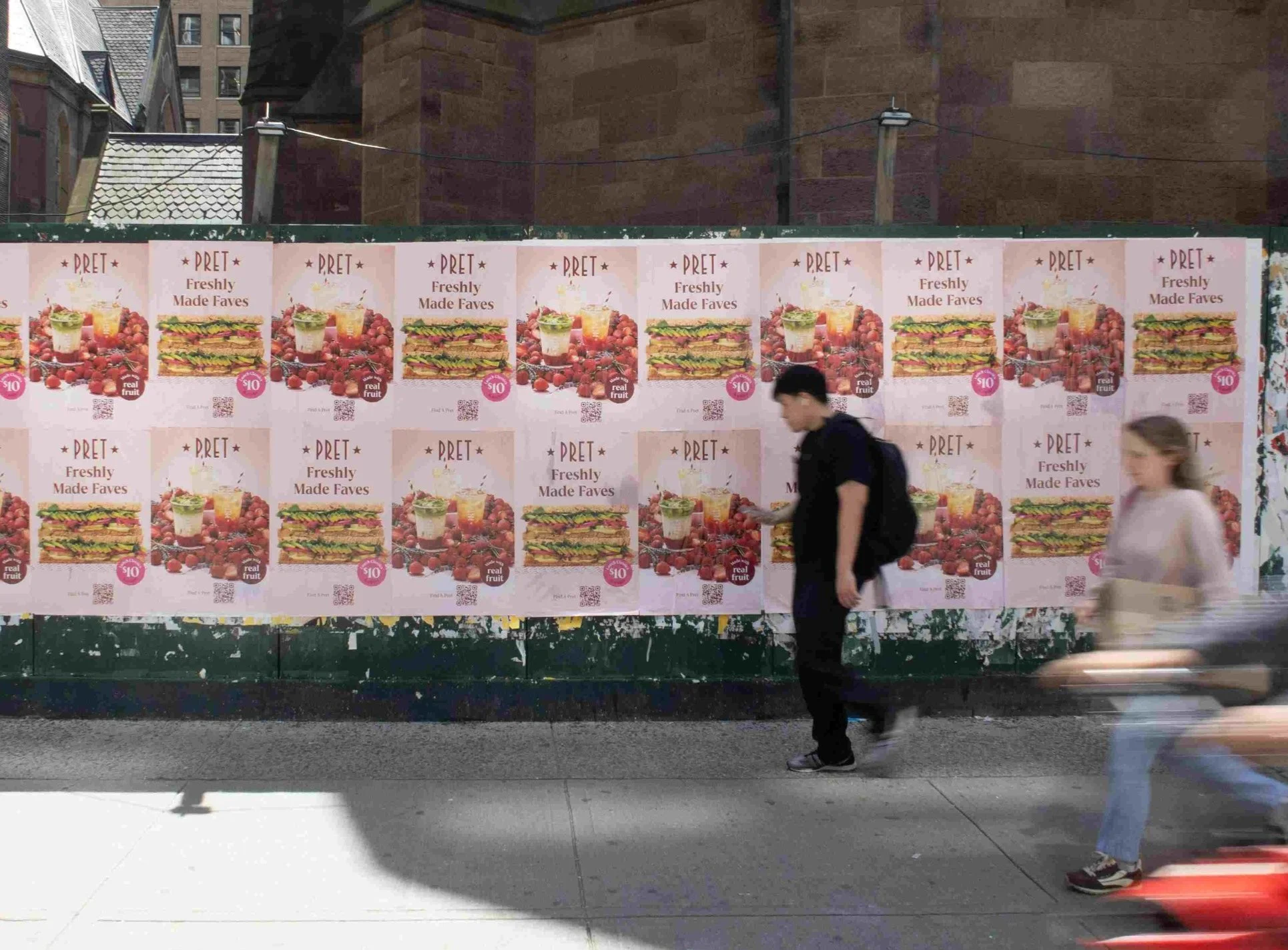 Street scene with a wall of posters advertising pre-made food, featuring sandwiches, fruit, and a drink, with two pedestrians walking past, one with a backpack and the other with a small bag.