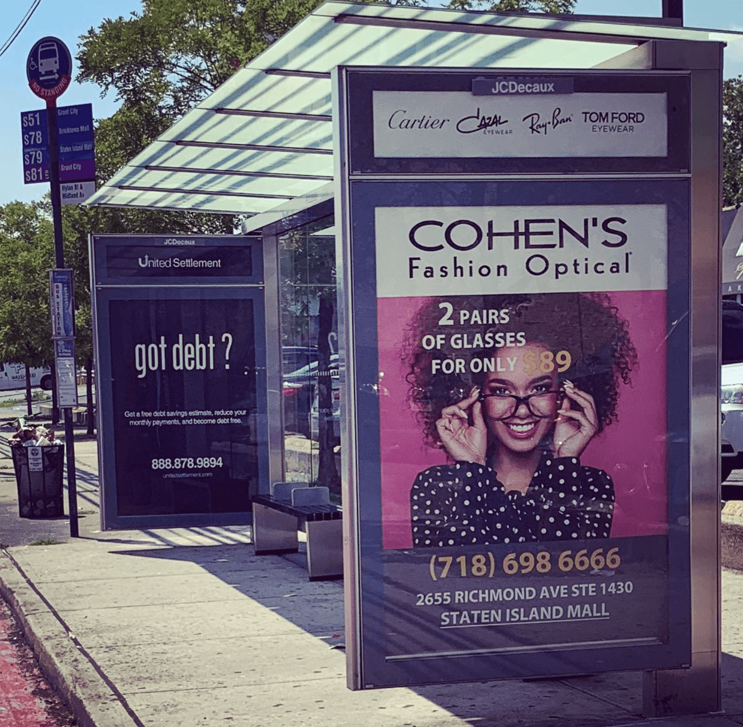 Bus stop shelter with two advertisements: one for Cohen's Fashion Optical promoting 2 pairs of glasses for $89, featuring a woman with curly hair and glasses, and another for United Settlement offering free debt savings estimates, with a phone number