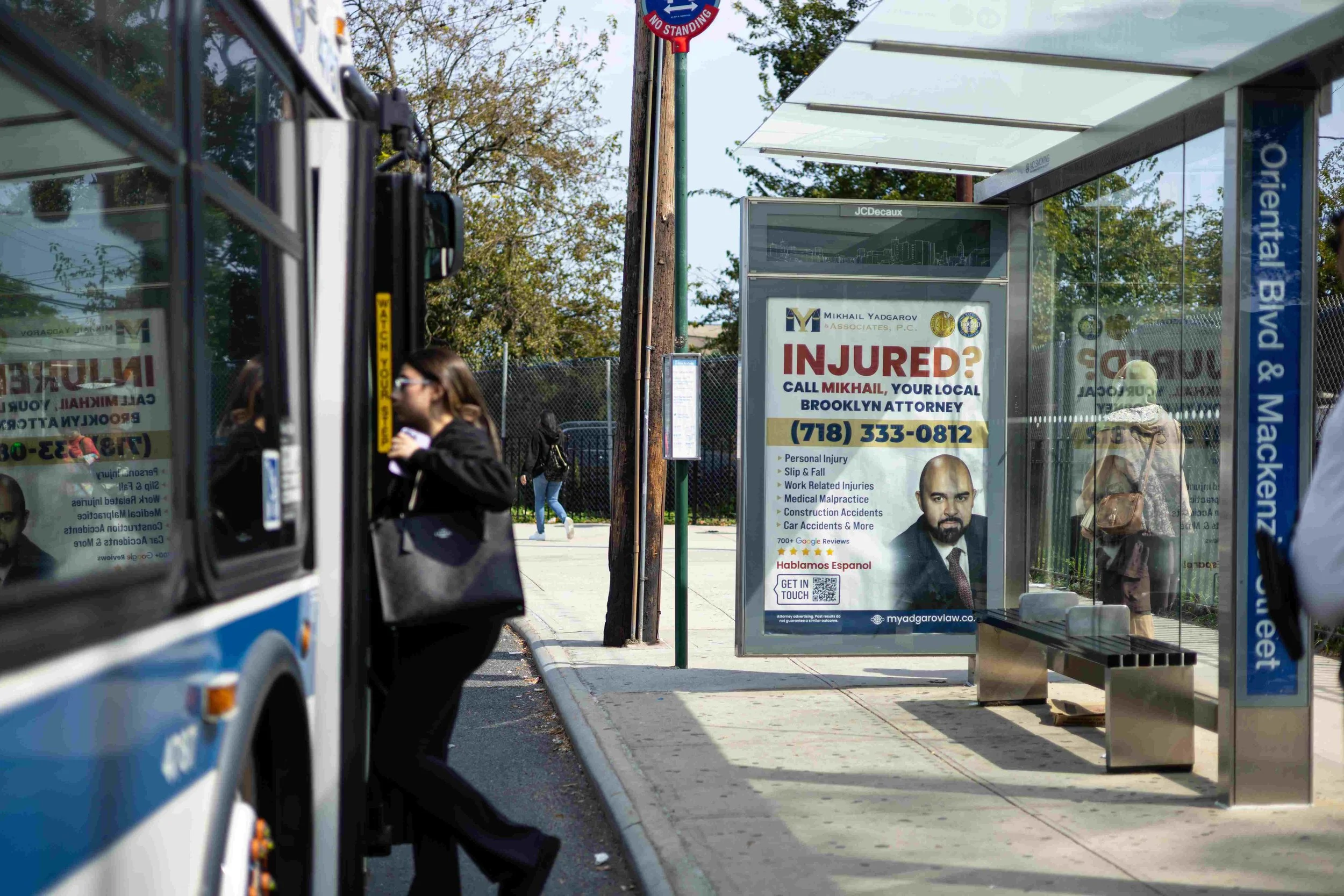 A woman is boarding a city bus at a bus stop in Brooklyn, New York. The bus stop has advertisements, including one for a personal injury attorney. There are other pedestrians in the background, and the bus has a blue and white color scheme.