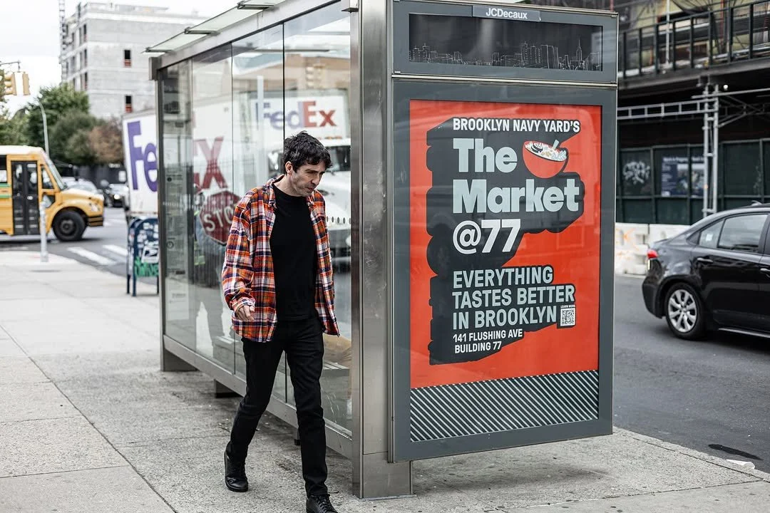 A man in a black shirt and plaid jacket walks past a bus stop with a large red and black advertisement for Brooklyn Navy Yard's The Market @77, which mentions that everything tastes better in Brooklyn and provides an address at 141 Flushing Ave, Buil