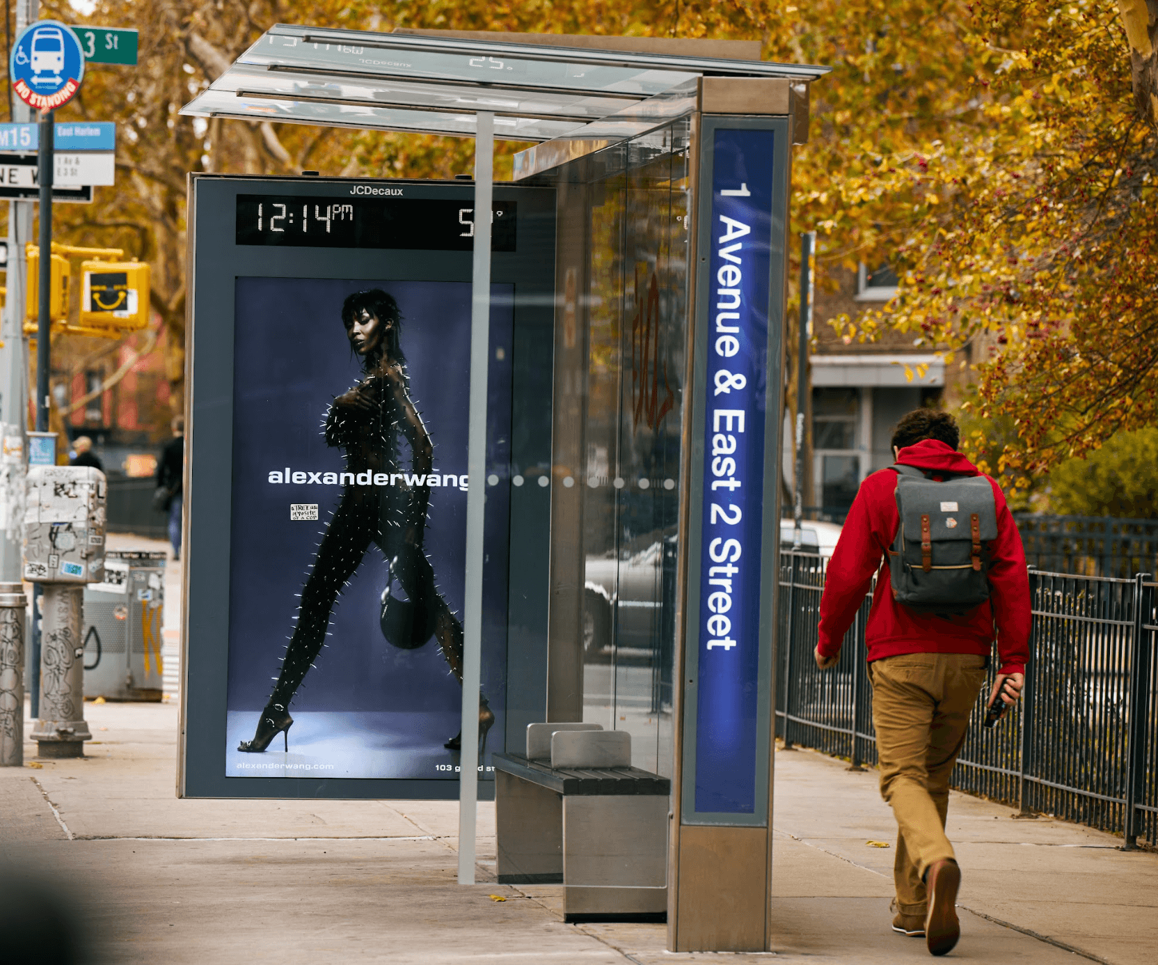 A person walking along a city sidewalk near a bus stop shelter with an advertisement for Alexander Wang, in an autumn setting with trees showcasing fall foliage.