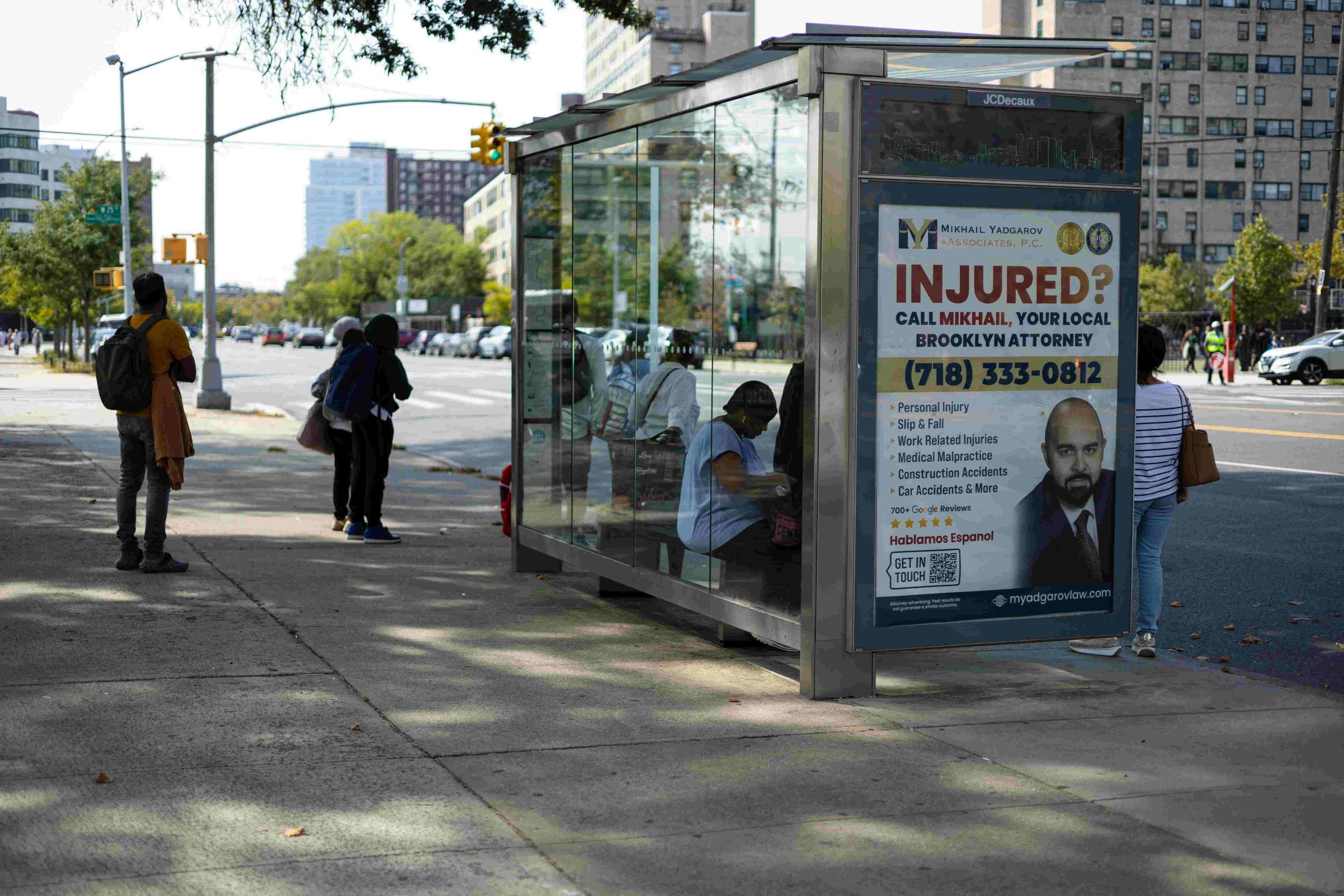 People waiting at a bus stop in an urban area with tall buildings, trees, and cars on the street. There is an advertisement on the bus shelter for a Brooklyn attorney, featuring a bald man in a suit, and contact information.