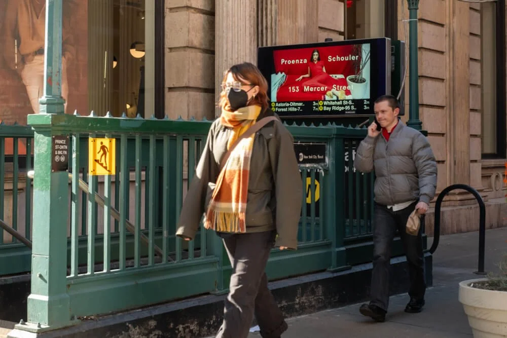 A woman wearing a face mask and a scarf walks past a subway entrance, while a man in a gray jacket talks on his phone nearby on a city street.