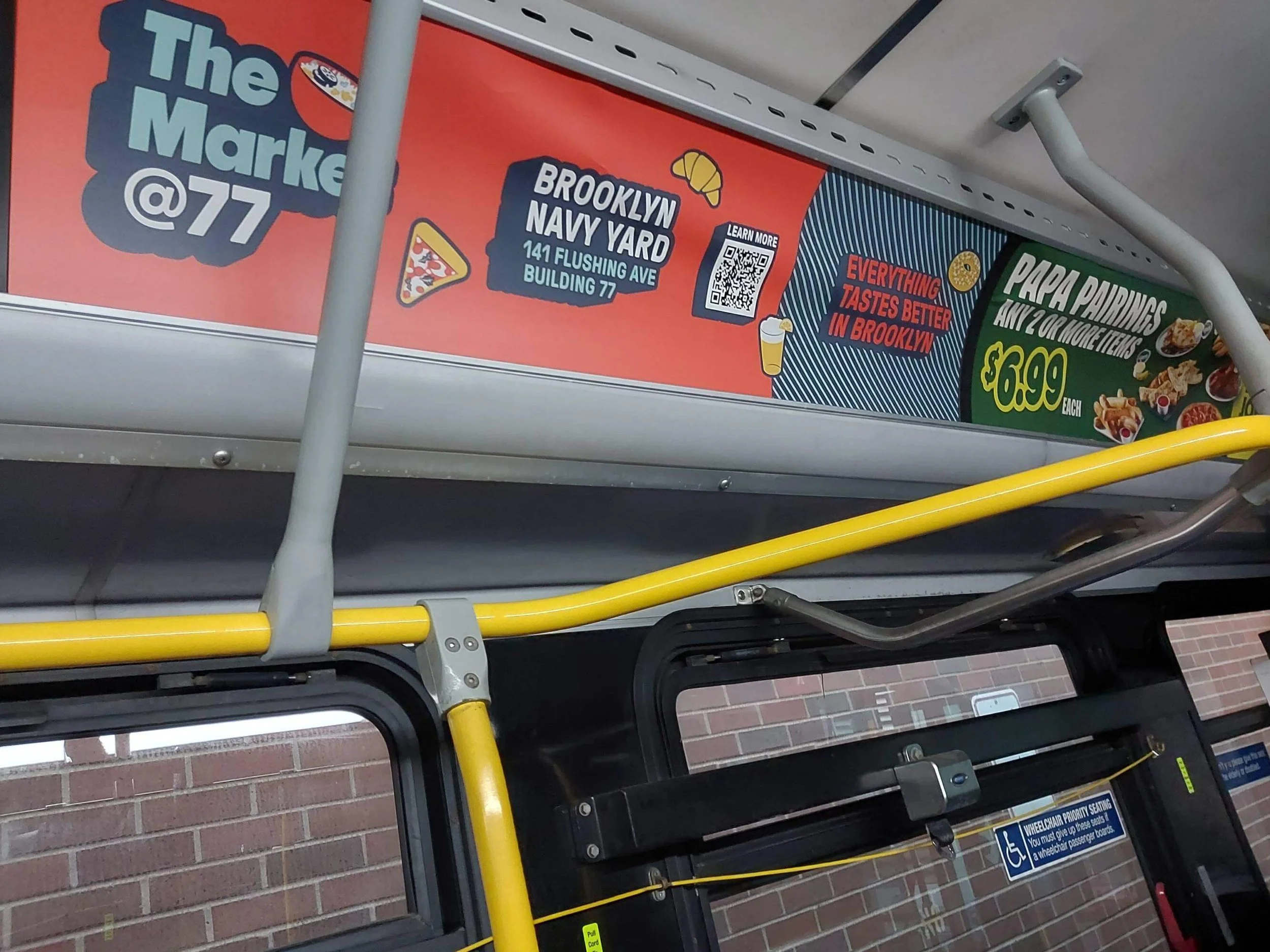 Advertisement on a public transit bus ceiling board advertising Brooklyn Navy Yard with address 141 Flushing Avenue, Building 77. The ad mentions pizza and toppings, services for Papa Raining with two or more items for $6.99, and includes colorful gr