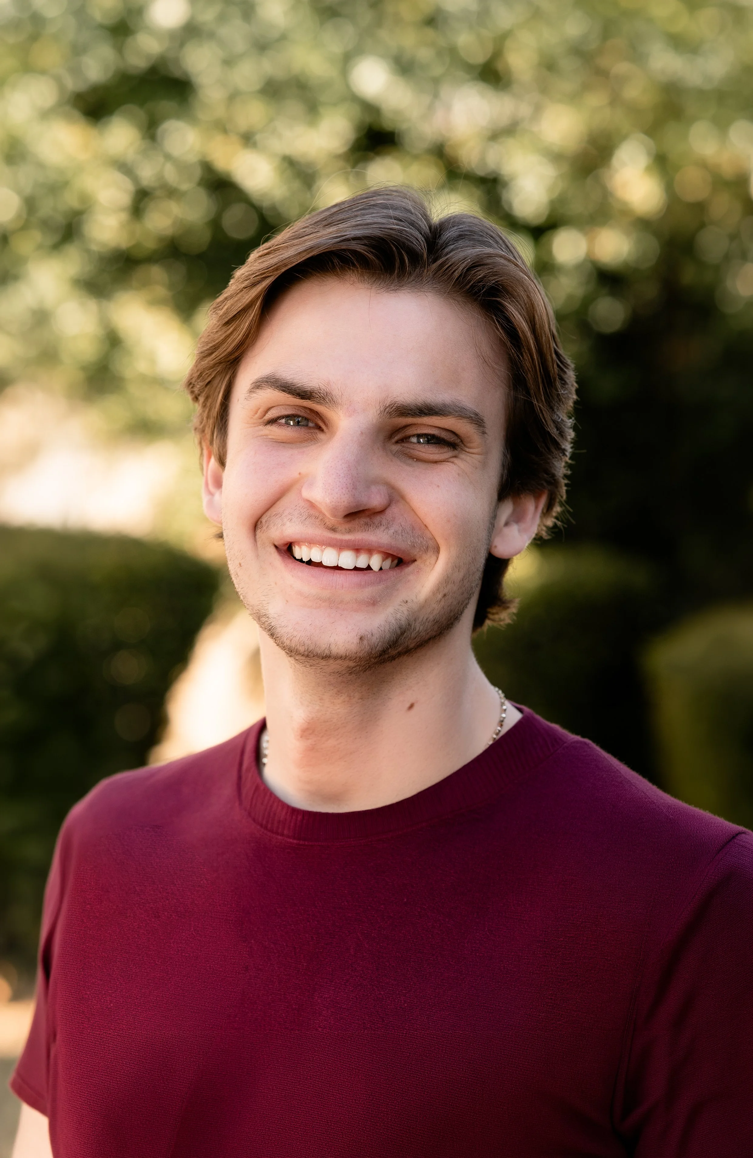 Young man smiling outdoors in front of green leafy background, wearing a black and gray checkered shirt.