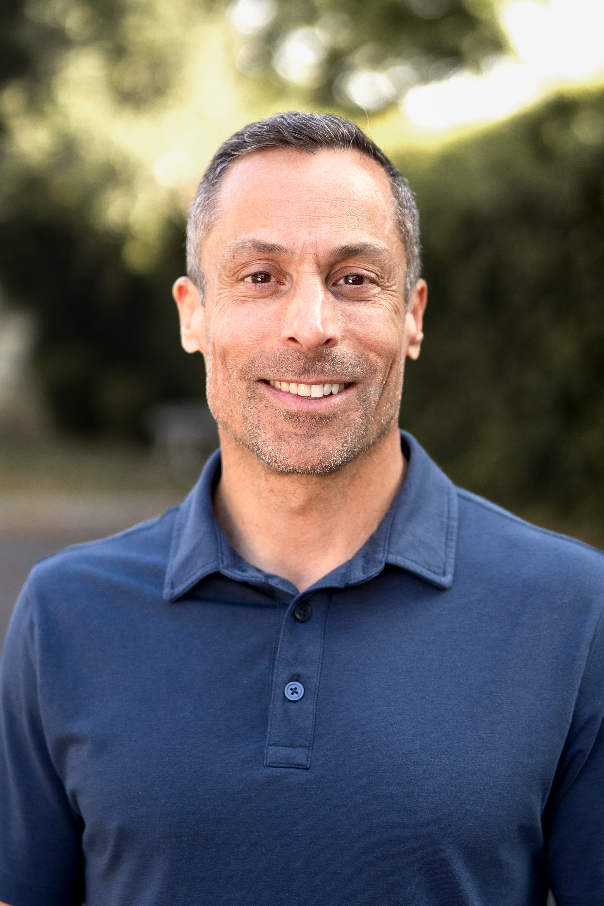A middle-aged man with short dark hair and a slight beard, smiling, wearing a blue polo shirt, outdoors with blurred greenery in the background.