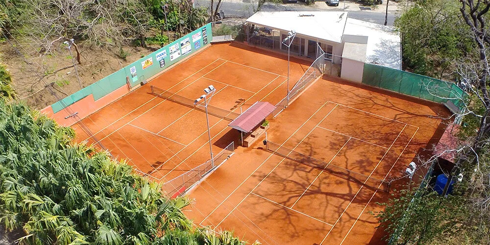 A view of an outdoor clay tennis court with multiple courts, surrounded by green fencing and trees, and a small building at the back.