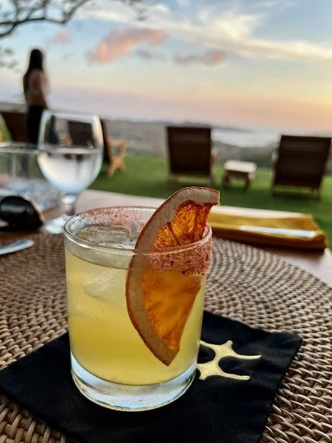 A glass of yellow cocktail garnished with a dried orange slice on a black napkin, on a woven table outdoors with a beach view and sitting area in the background.