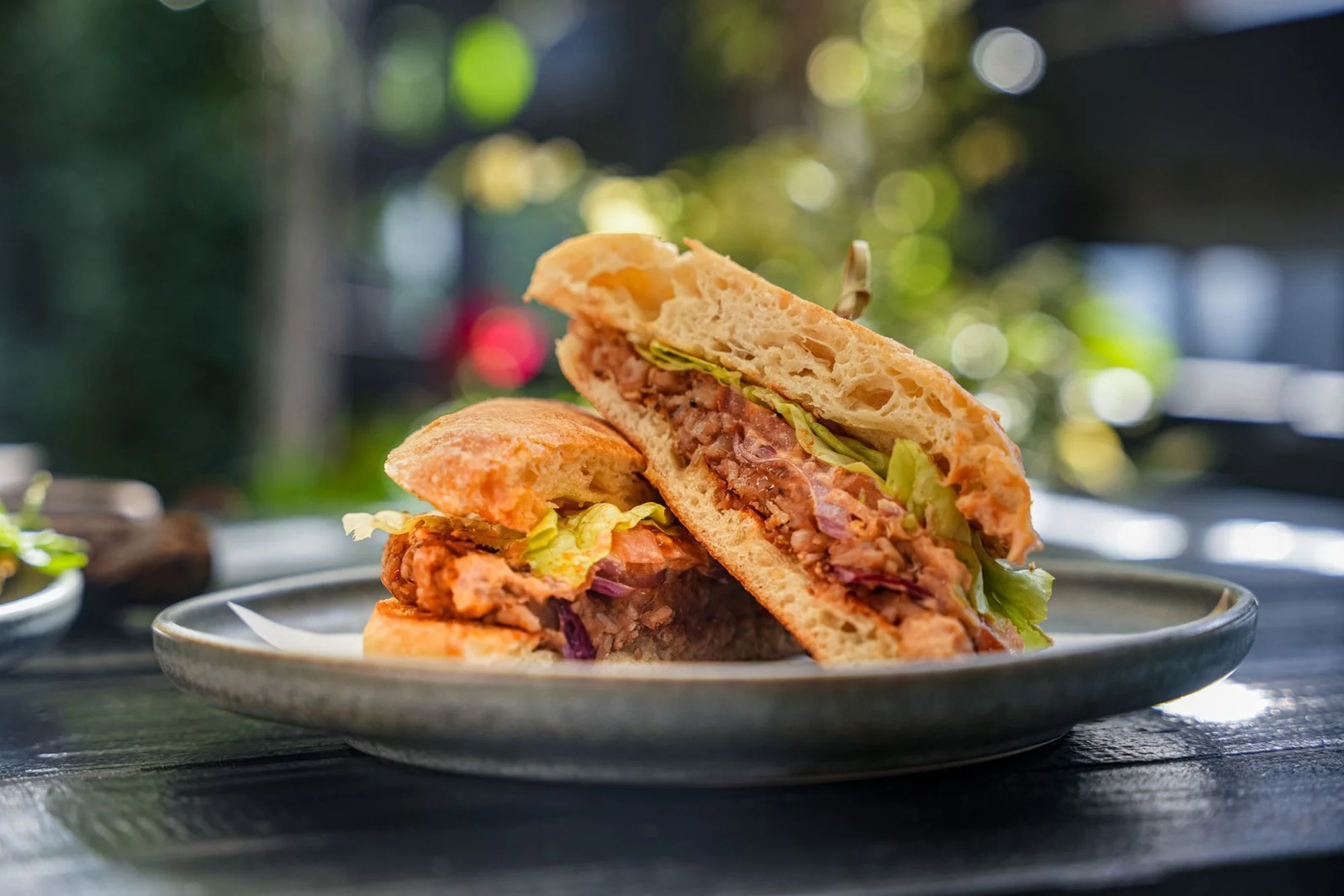 A sandwich with lettuce, tomato, and meat on a ceramic plate outside with a blurred background of trees and sunlight.