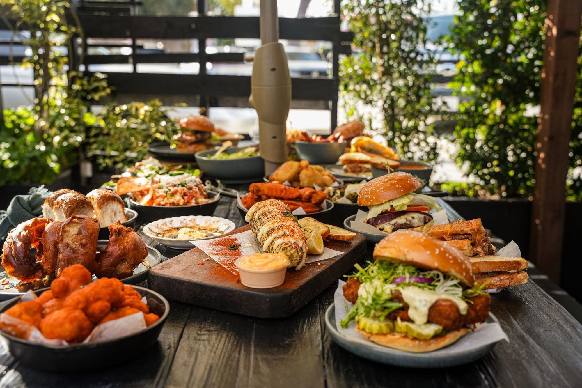 Outdoor table filled with various dishes including burgers, fried chicken, sushi, and sides, set in a patio area with greenery and wooden fencing.