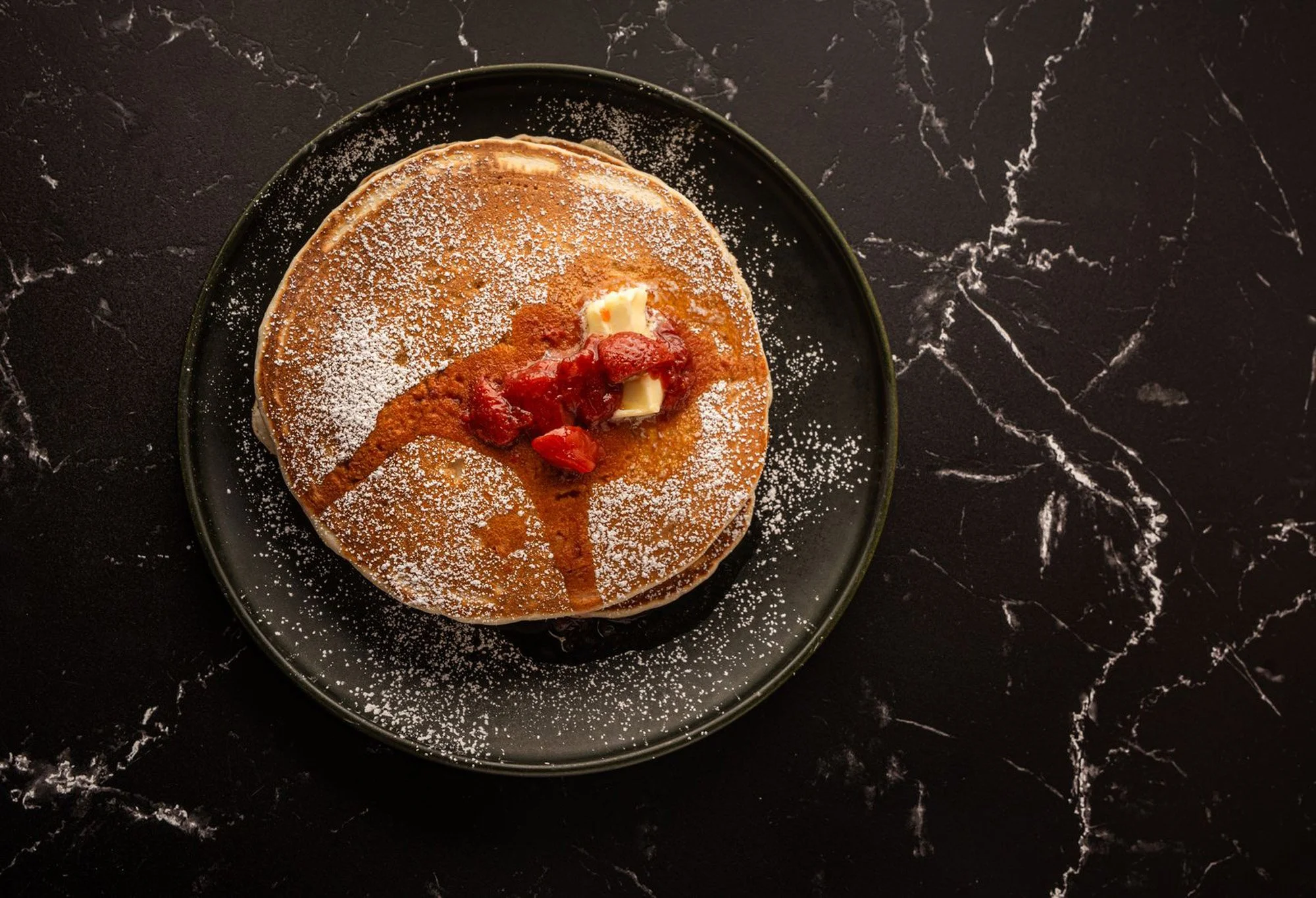 A stack of pancakes topped with butter, strawberry sauce, and powdered sugar on a black plate placed on a black marble surface.