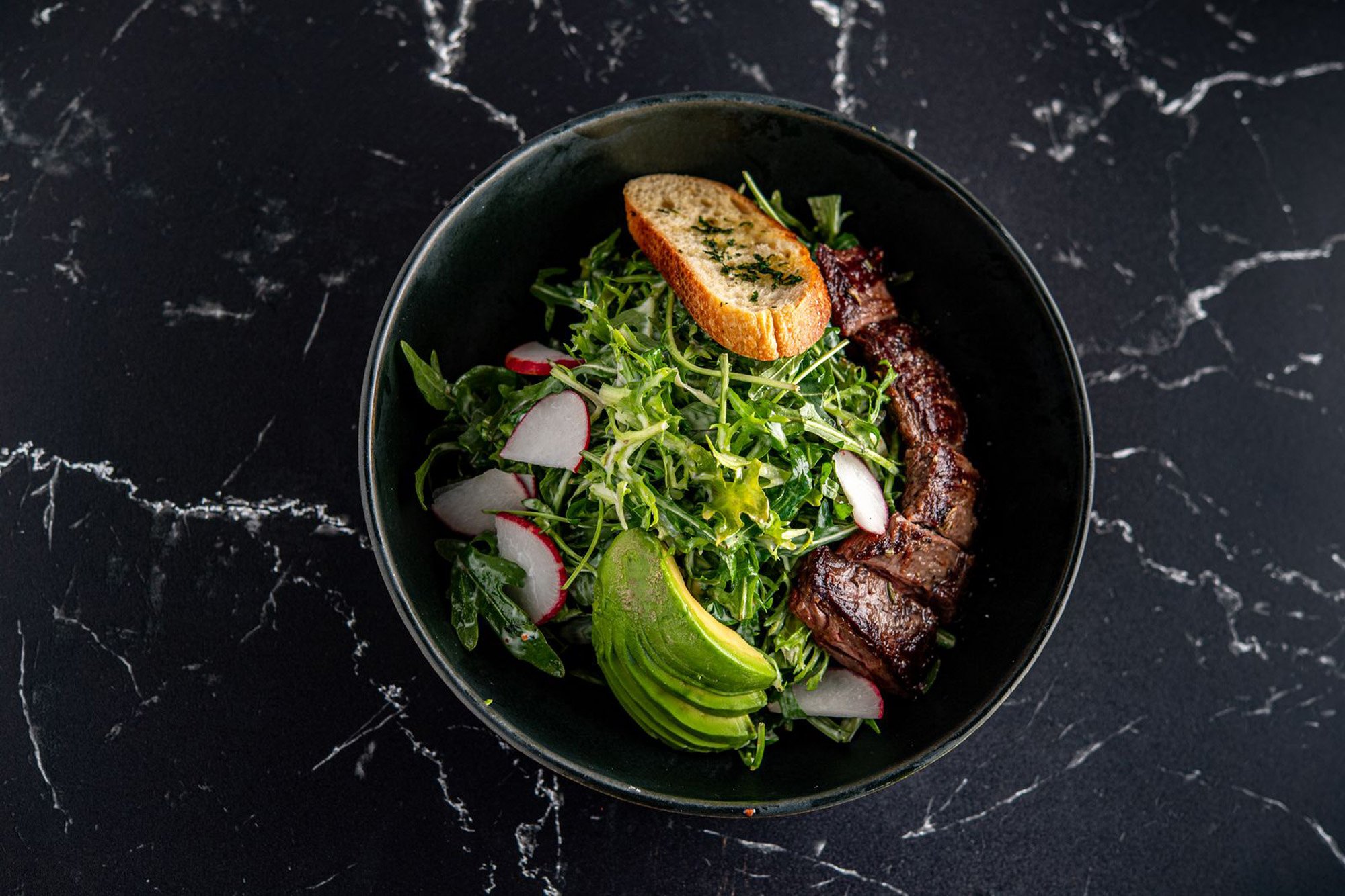 A bowl containing a fresh salad with radish slices, avocado slices, leafy greens, a piece of toasted garlic bread, and grilled beef.
