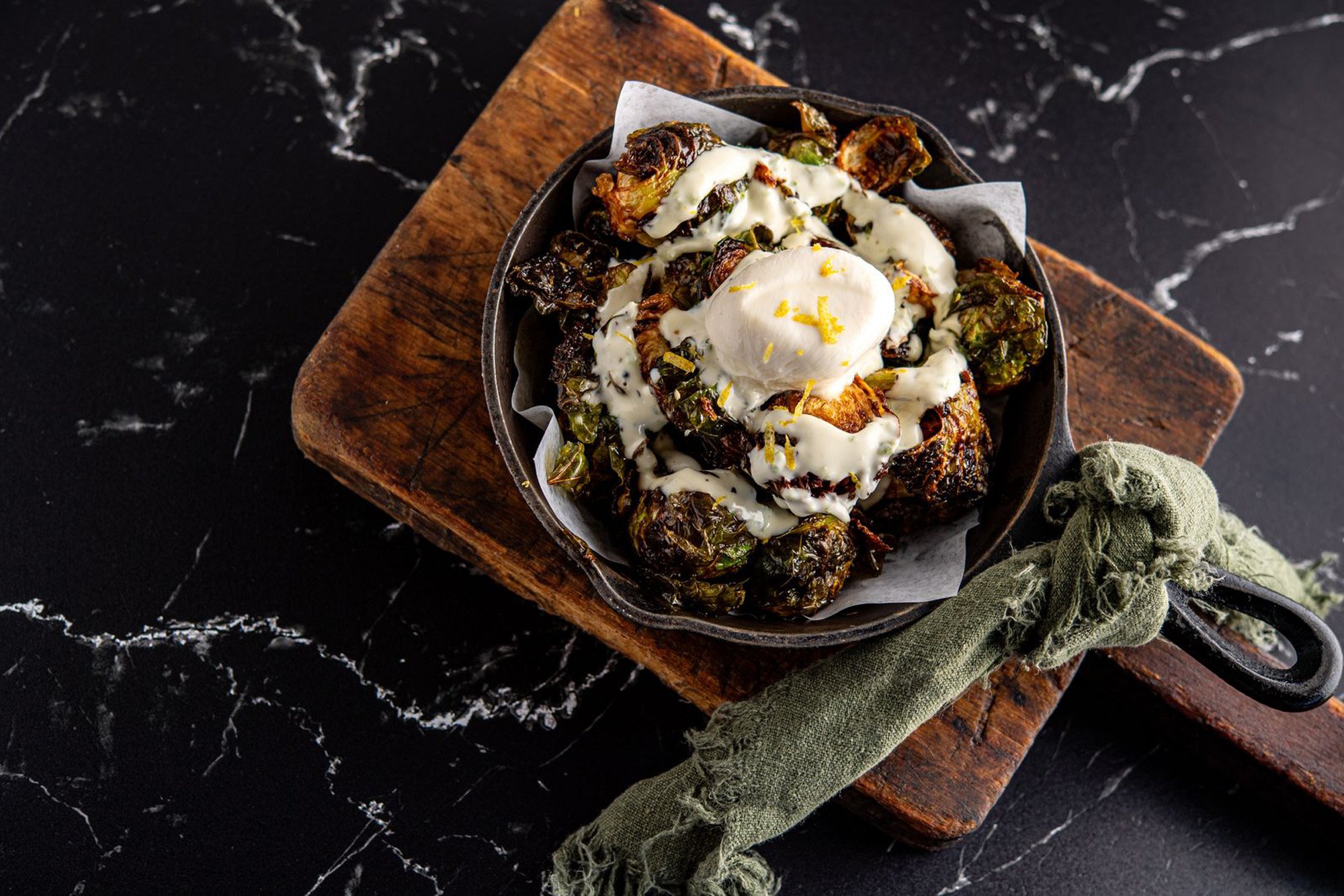 A black cast iron skillet filled with roasted brussels sprouts topped with a dollop of sour cream and lemon zest, placed on a wooden cutting board on a black marble surface.