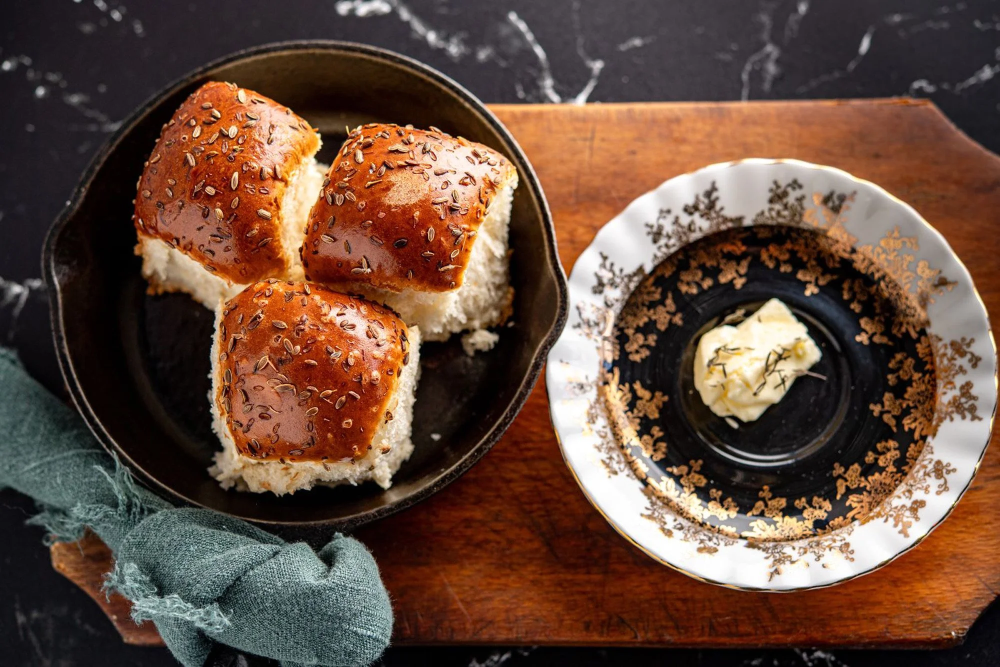Three pieces of bread with shiny, sesame and cumin seed topping on a black plate, accompanied by a small dish of butter or margarine on a decorative white plate with gold patterns, placed on a wooden tray.