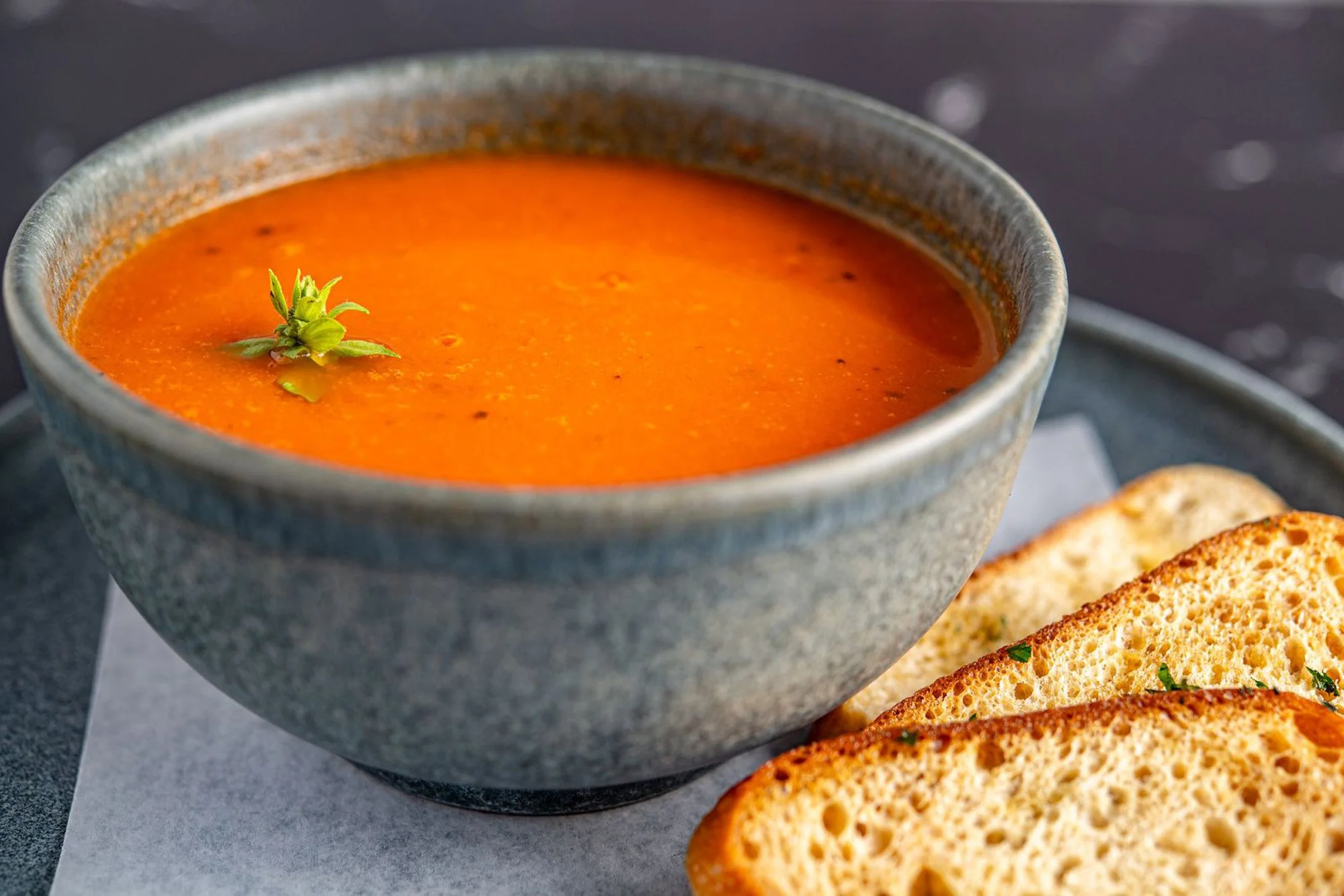 A bowl of tomato soup garnished with a small sprig of fresh herbs, served with slices of toasted bread on a tray.