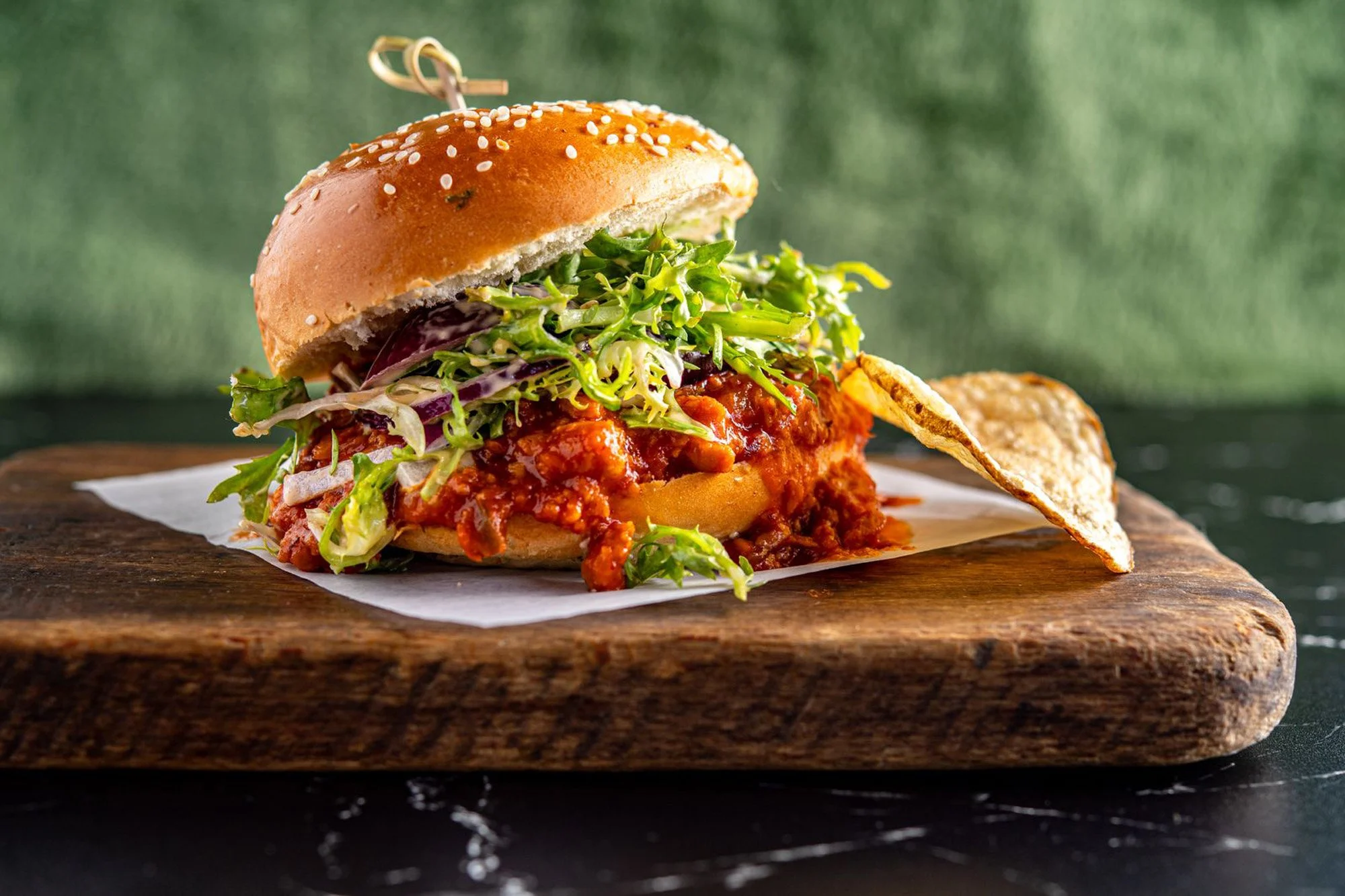 Close-up of a pulled pork sandwich with lettuce, barbecue sauce, and pickles in a sesame seed bun, served with a potato chip on a wooden cutting board.