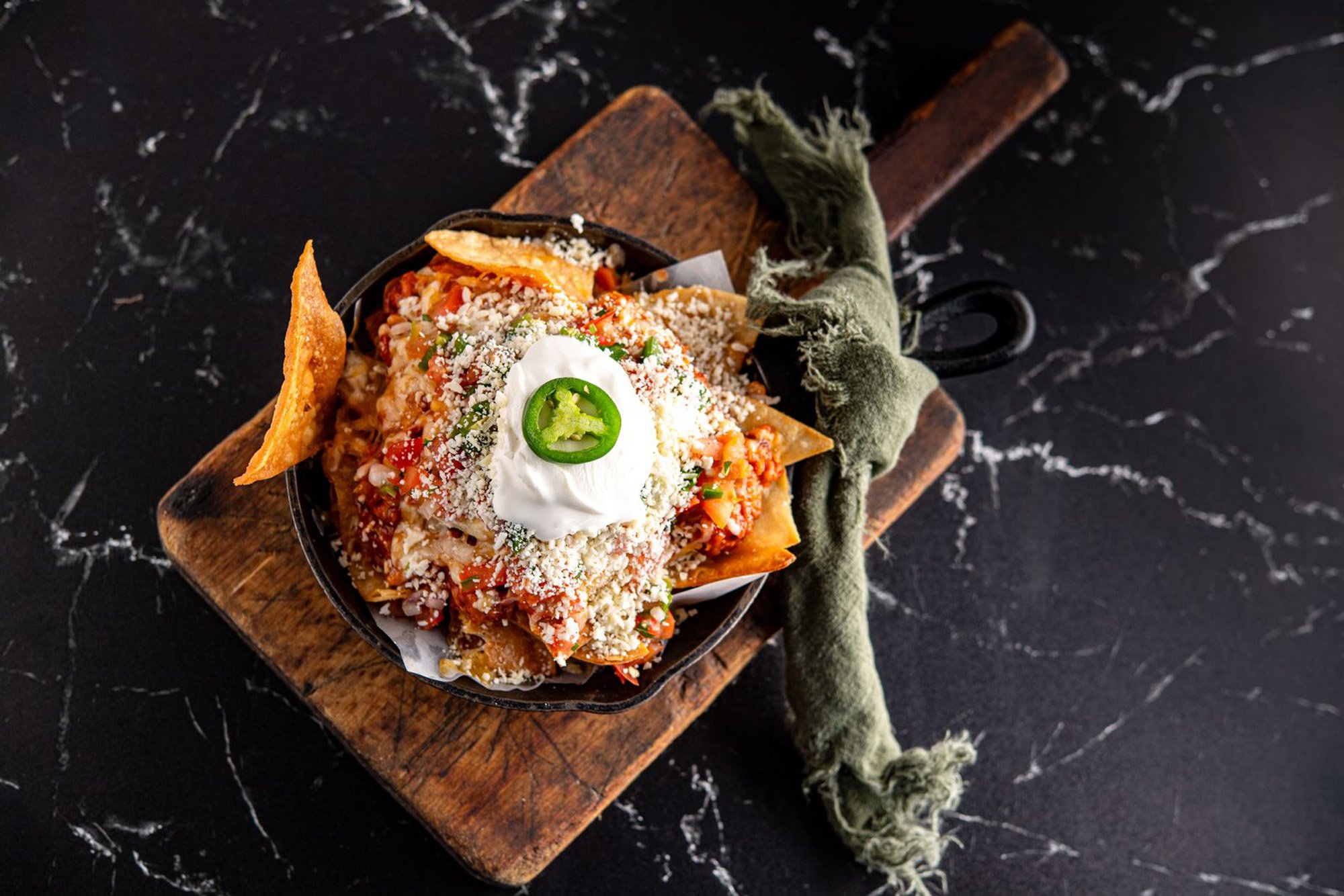 Top-down view of a serving of nachos with melted cheese, diced tomatoes, sour cream, sliced jalapeño, and shredded cheese, on a black skillet placed on a wooden board with a rustic cloth.