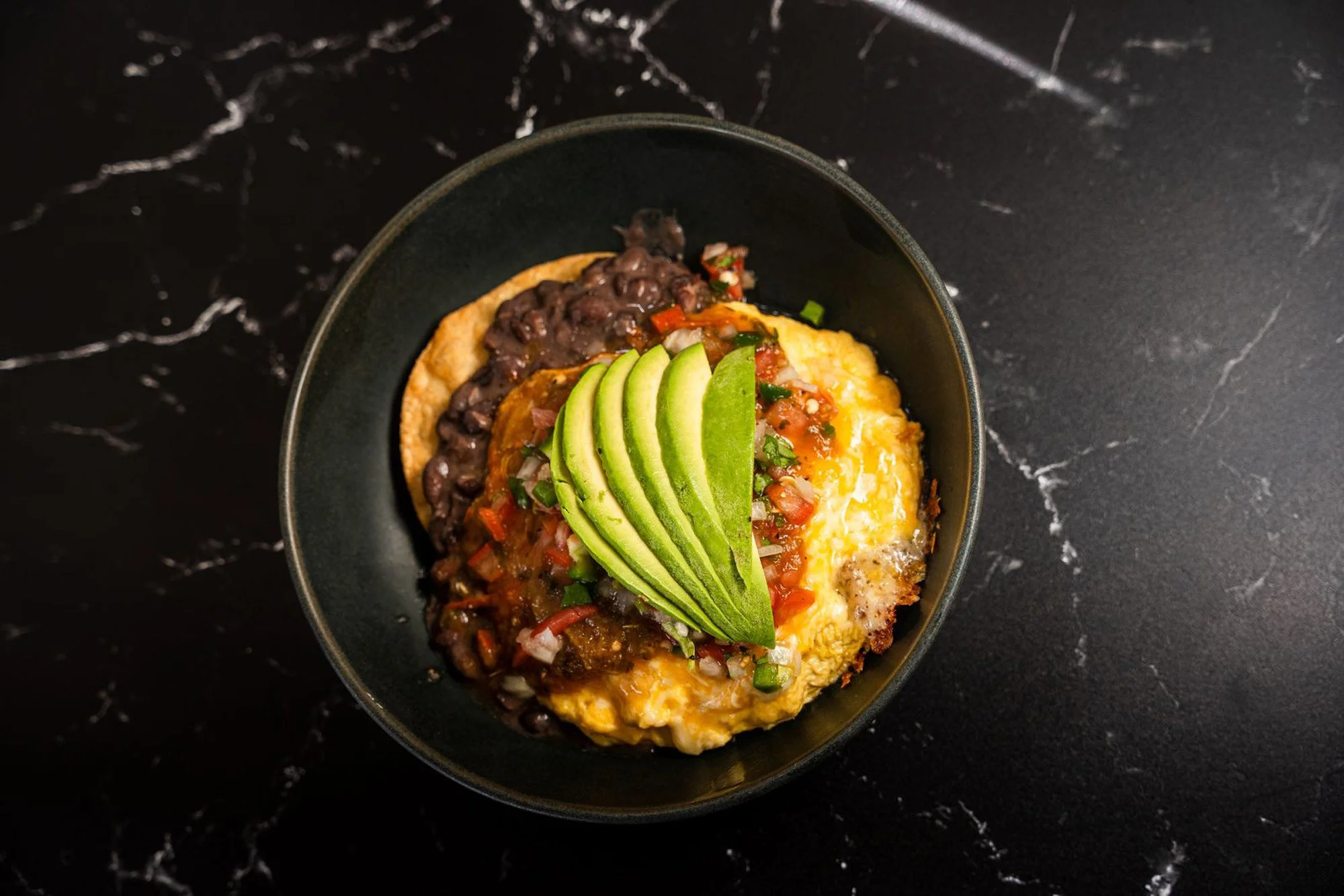A black bowl filled with a Mexican-style breakfast dish including scrambled eggs, black beans, sliced avocado, and a tomato-based salsa, placed on a dark marble surface.