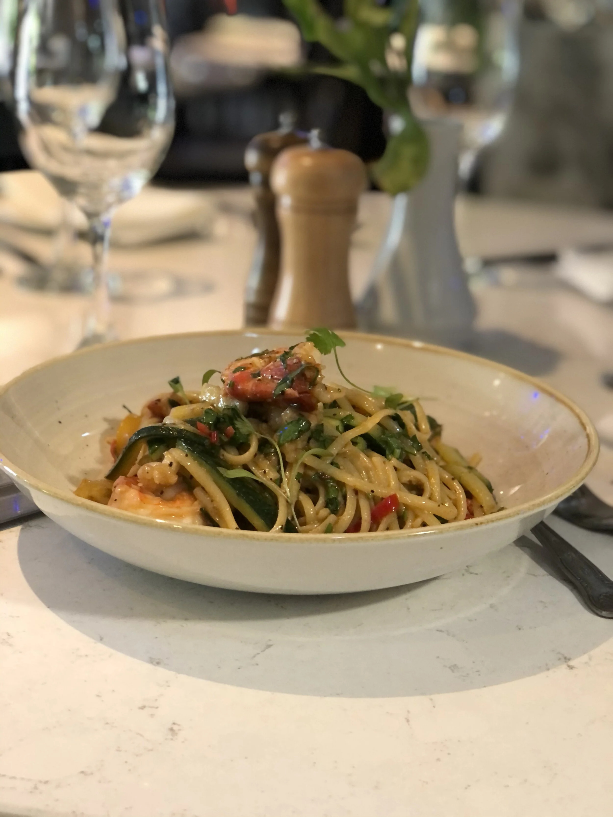 A bowl of Asian noodle dish with vegetables and seafood, garnished with herbs, placed on a white table in a restaurant setting.