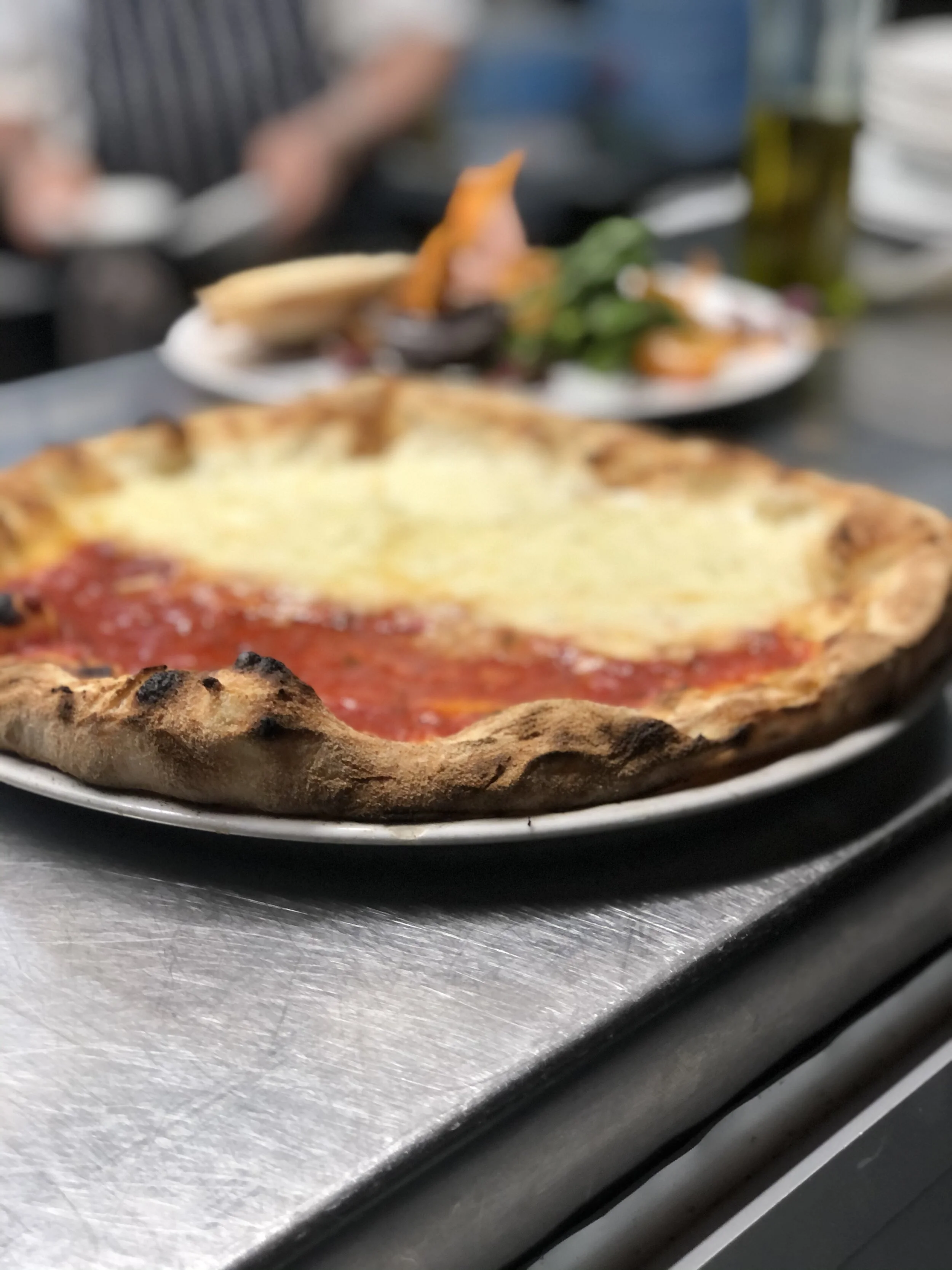 A close-up of a pizza with cheese and tomato sauce on a plate, with a blurred background of people and dishes at a restaurant.