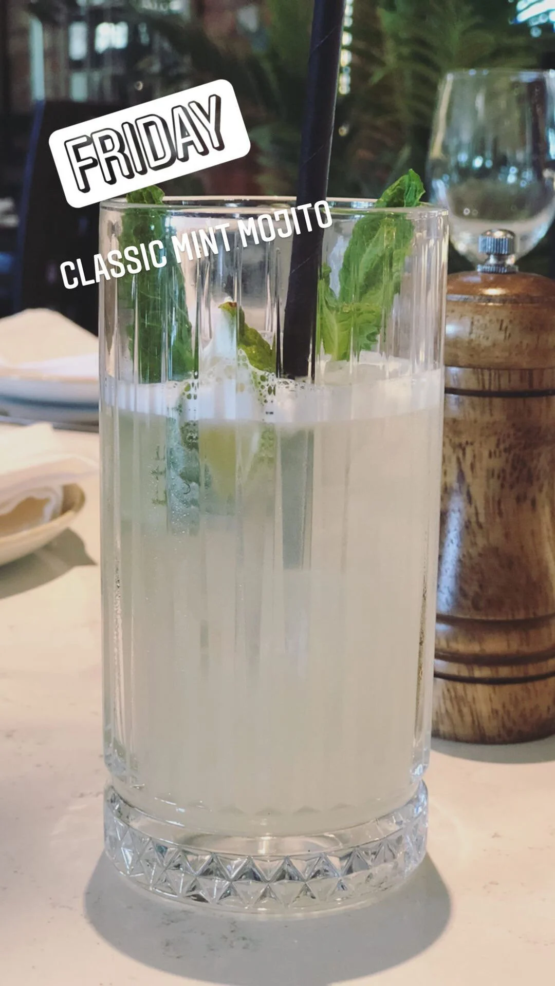 A tall glass of classic mint mojito with fresh mint leaves, lime, and a black straw on a table at a restaurant.