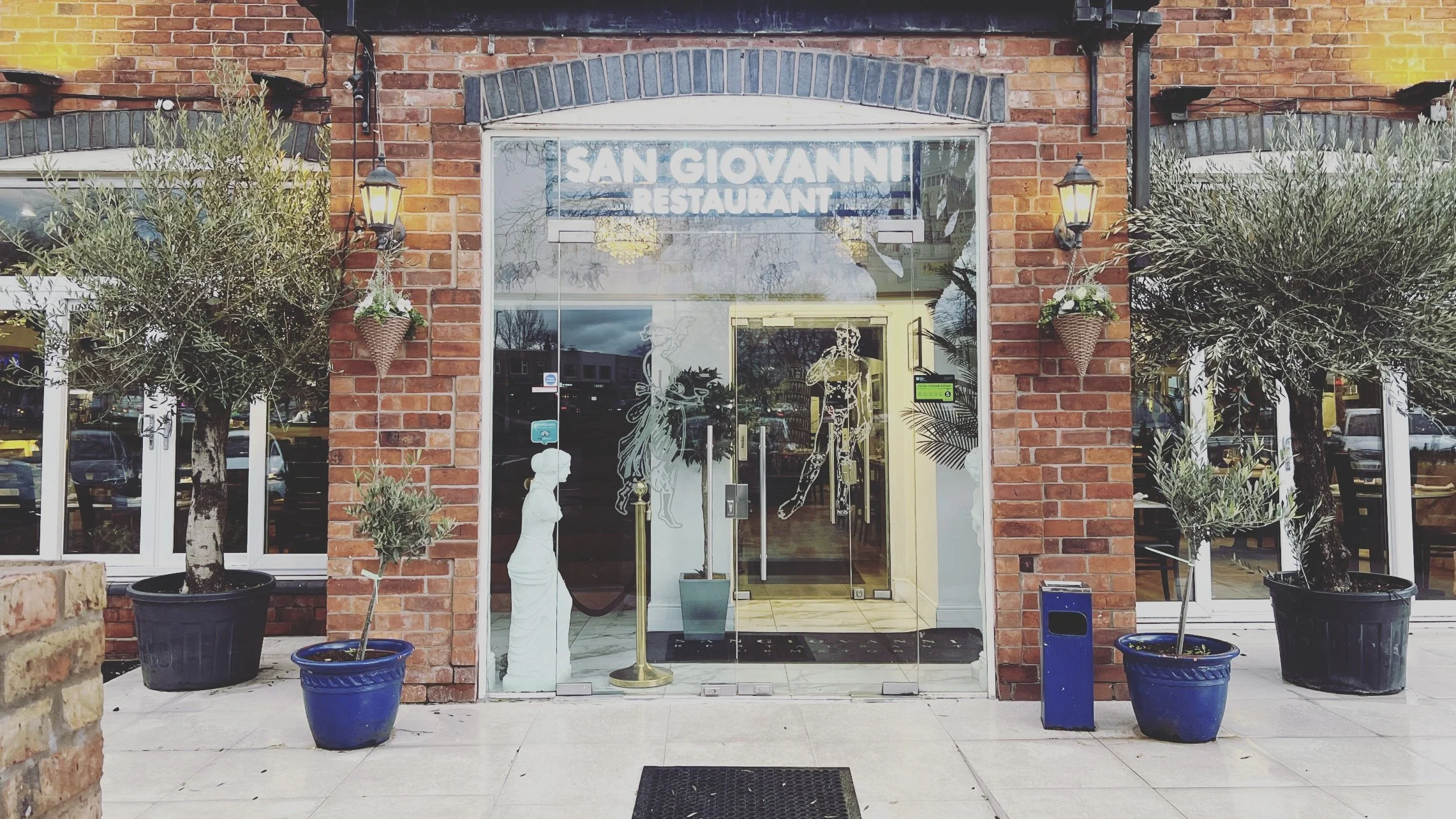 Exterior view of San Giovanni Restaurant with brick walls, glass door, potted olive trees, and decorative sculptures at entrance.
