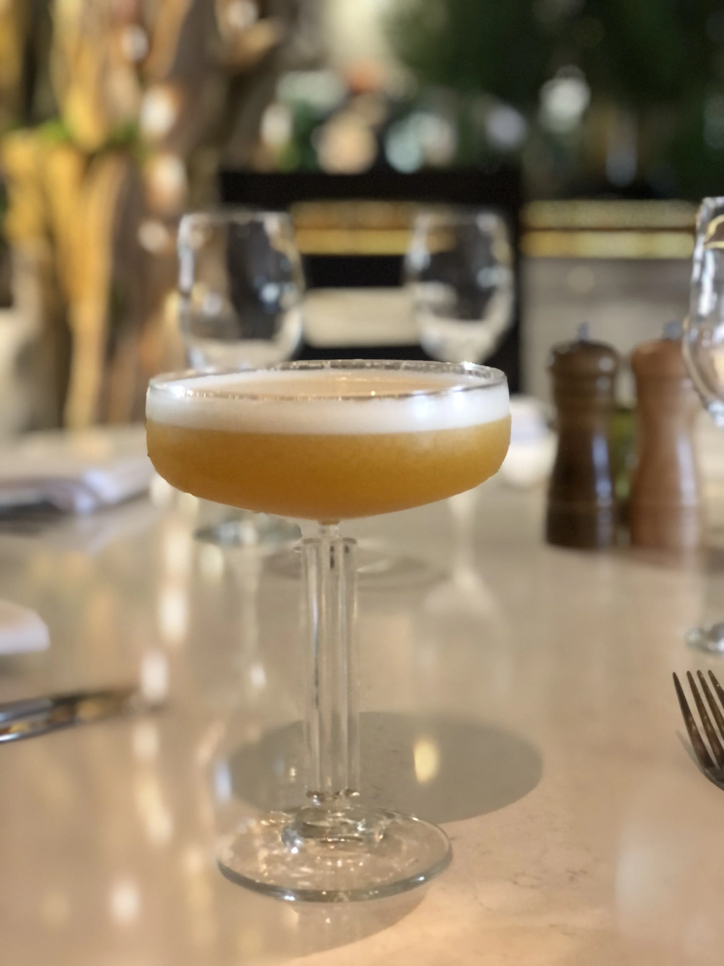 A cocktail in a coupe glass with a frothy top on a restaurant table, with utensils and salt and pepper shakers in the background.