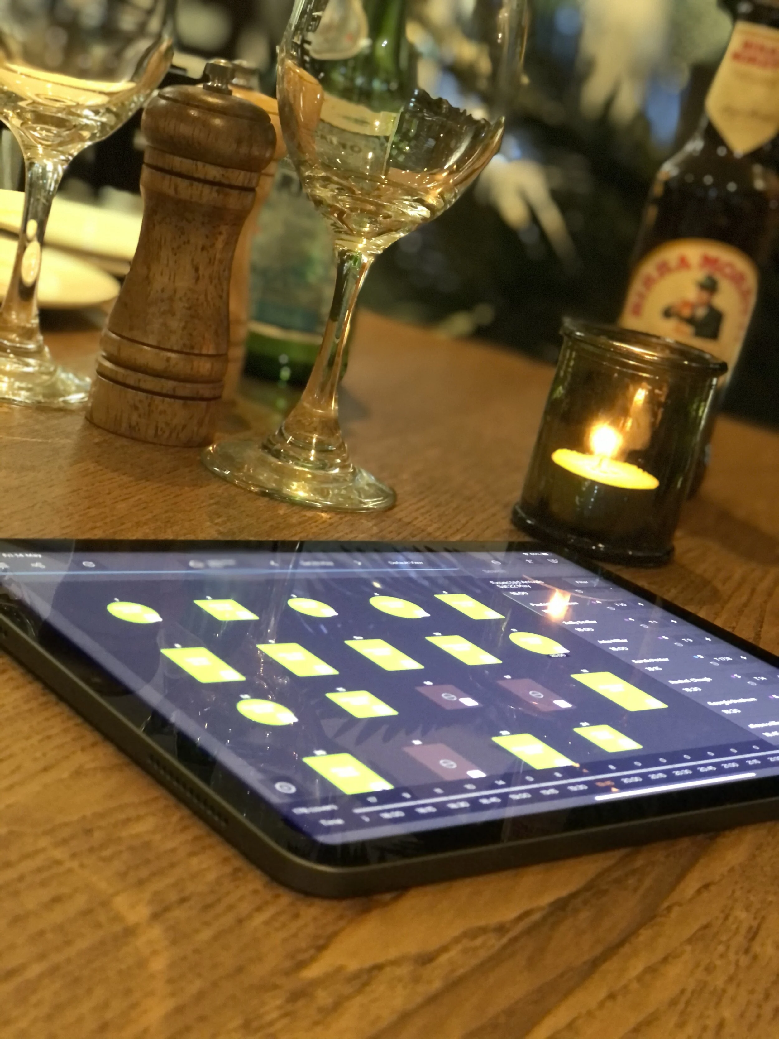 A smartphone on a wooden table displaying a music production app, with various yellow pads and controls. Behind the phone are empty wine glasses, a wooden pepper grinder, a lit candle in a glass holder, a green glass bottle, and a partially visible b