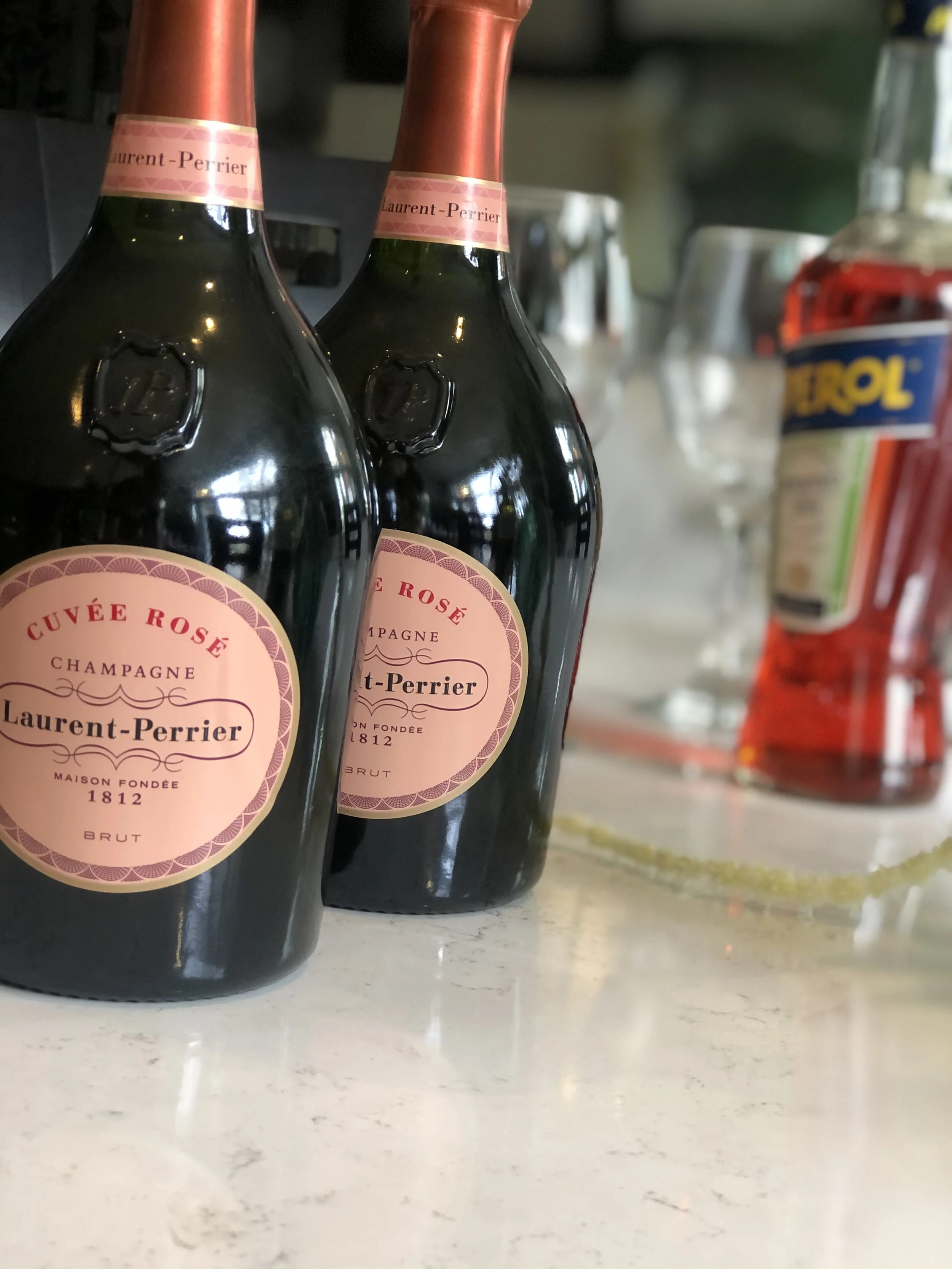 Two bottles of Laurent-Perrier Cuvée Rosé champagne with pink labels, placed on a white surface with a blurry background including a bottle of Trool spirits and other glassware.