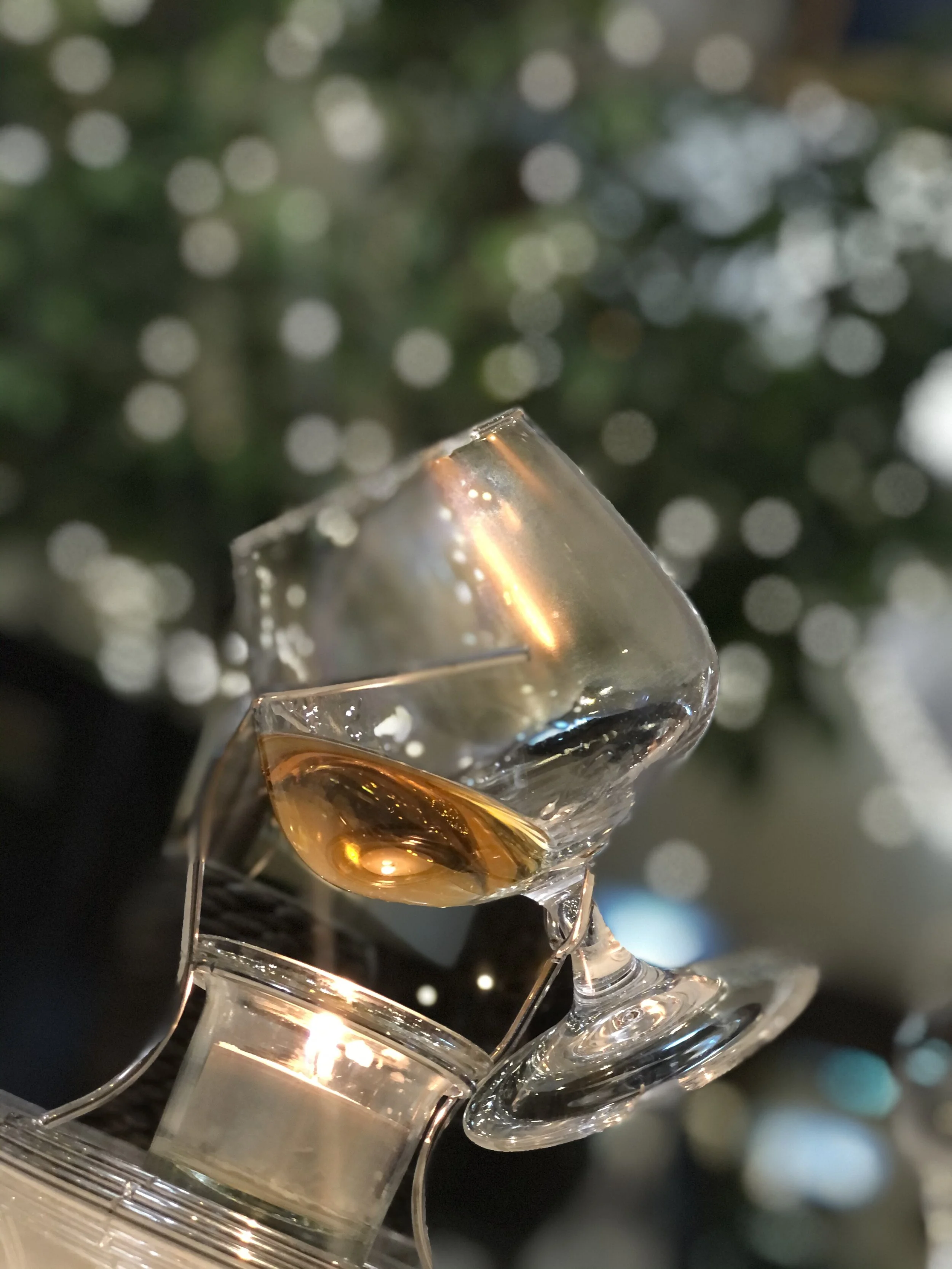 A close-up of a partially filled glass of whiskey with ice, reflecting warm light, with a blurred background of holiday lights.