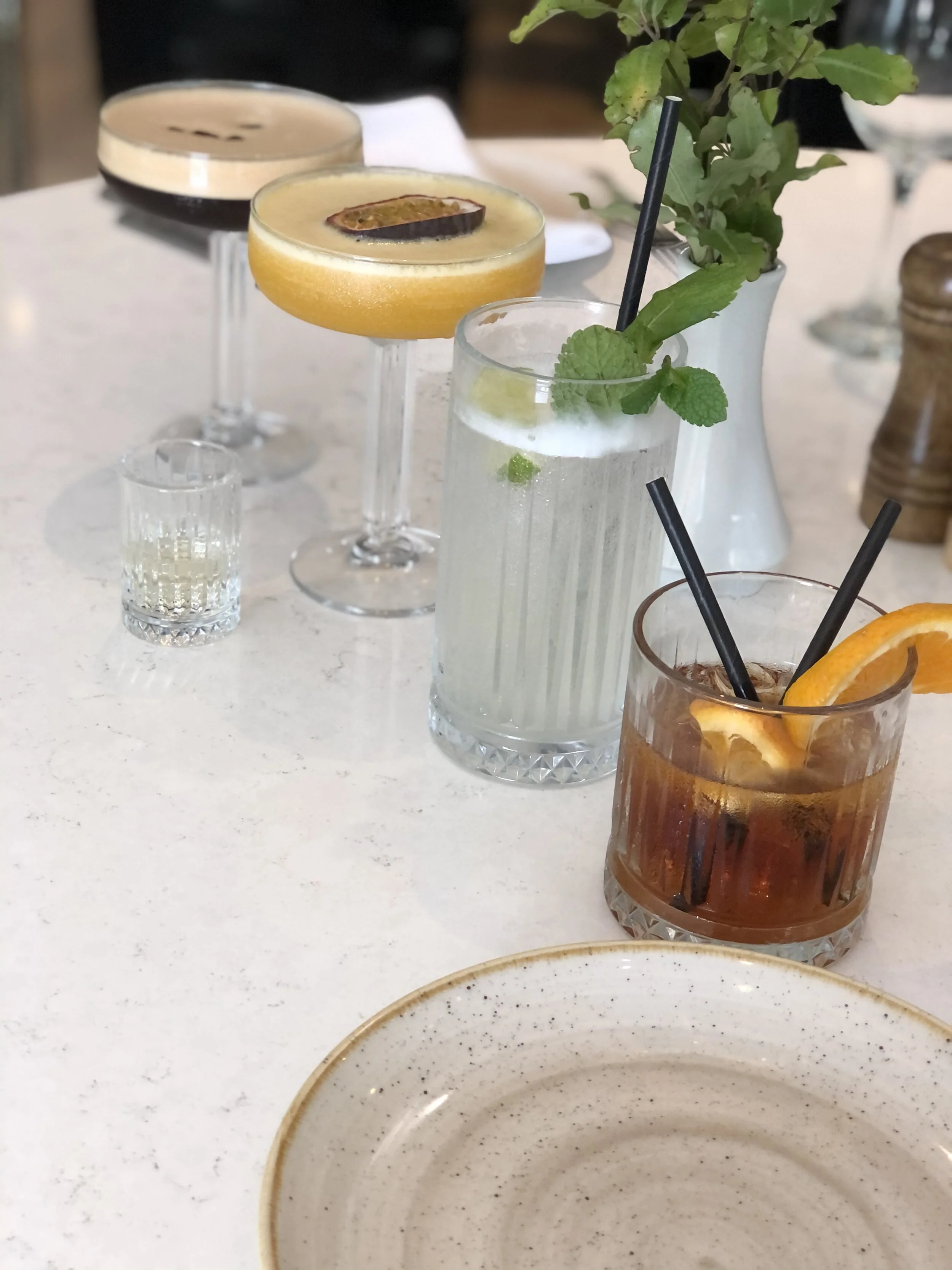 A row of various cocktails on a white table, including a dark cocktail in a coupe glass with a wooden garnish, a yellow cocktail with a slice of citrus fruit, a clear cocktail with mint leaves, and an amber-colored drink with an orange slice. There i