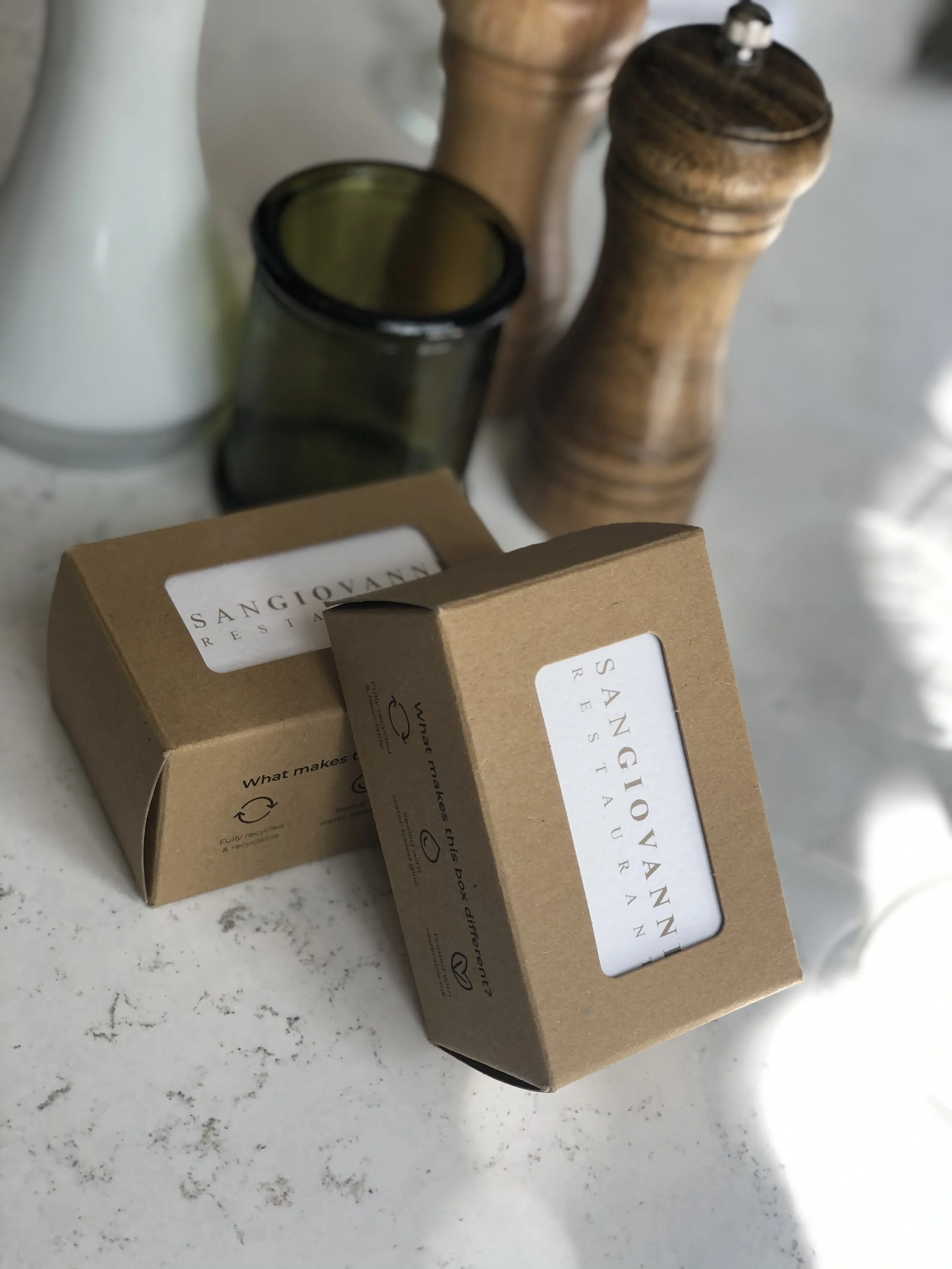 Two cardboard boxes labeled 'Sangiovanni Restaurant' on a white surface, with wooden and glass containers in the background.