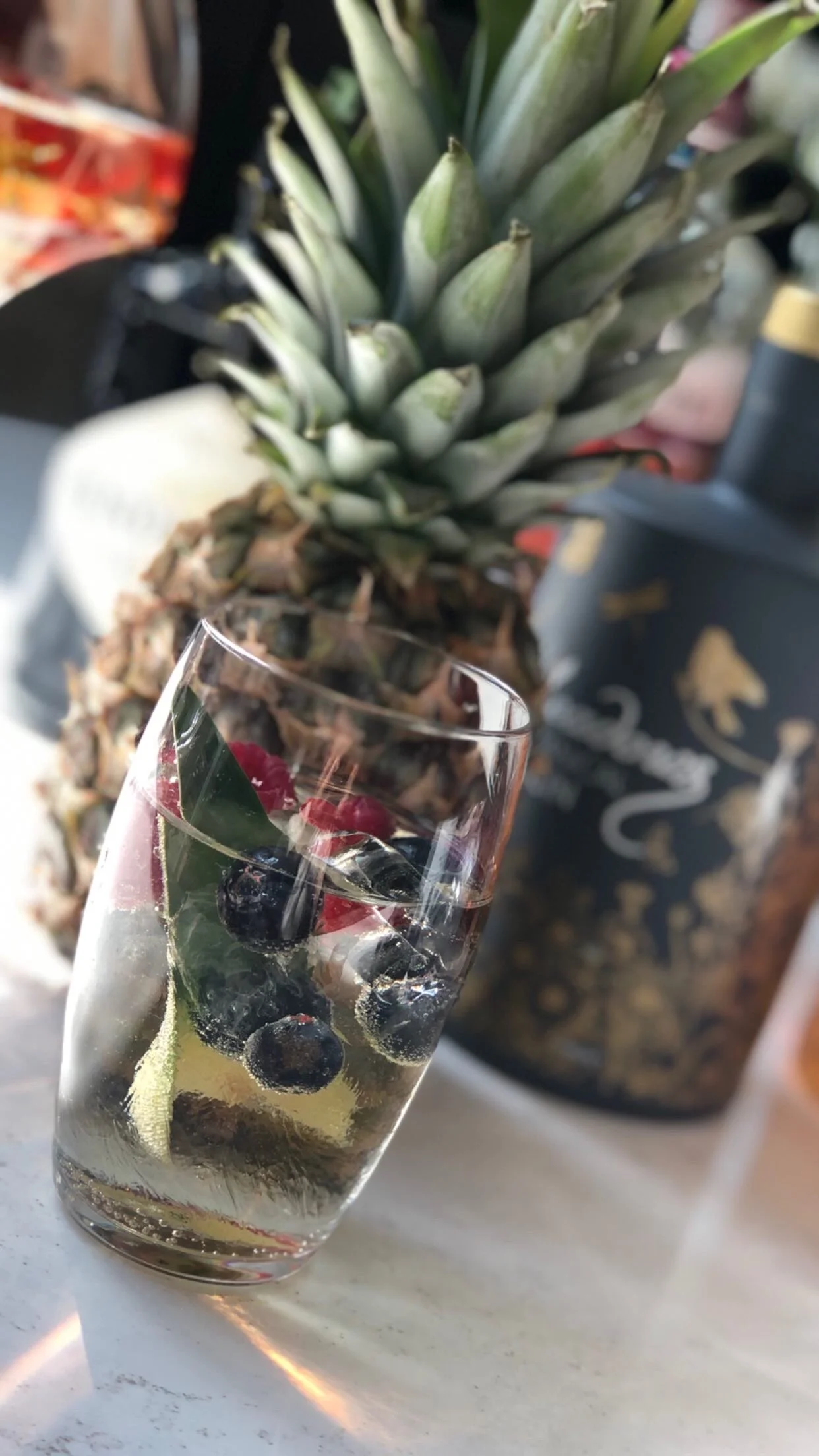 A glass of sparkling water with blueberries, raspberries, and a slice of lemon, placed on a white surface with a pineapple and a decorative planter in the background.