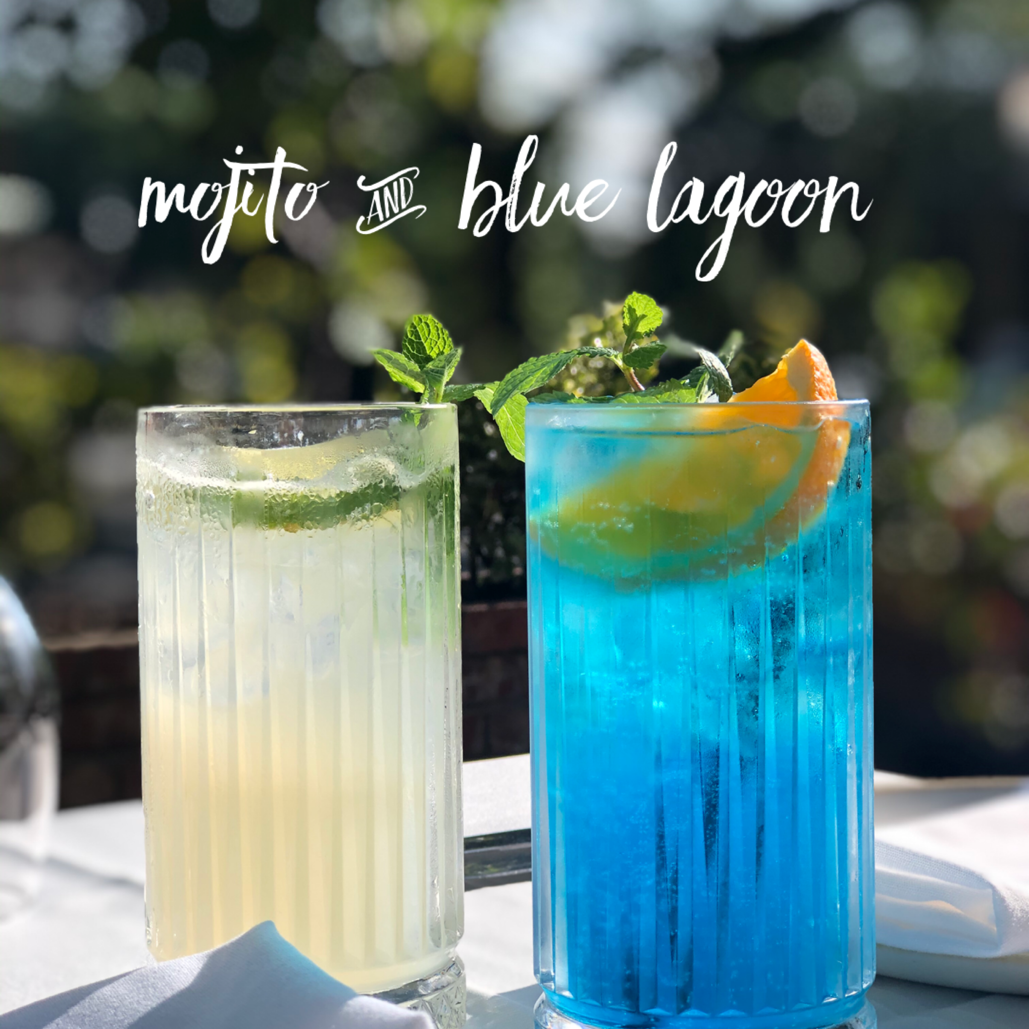 Two colorful cocktails, one yellow and one blue, garnished with lemon slices and mint leaves, outdoors on a white table.