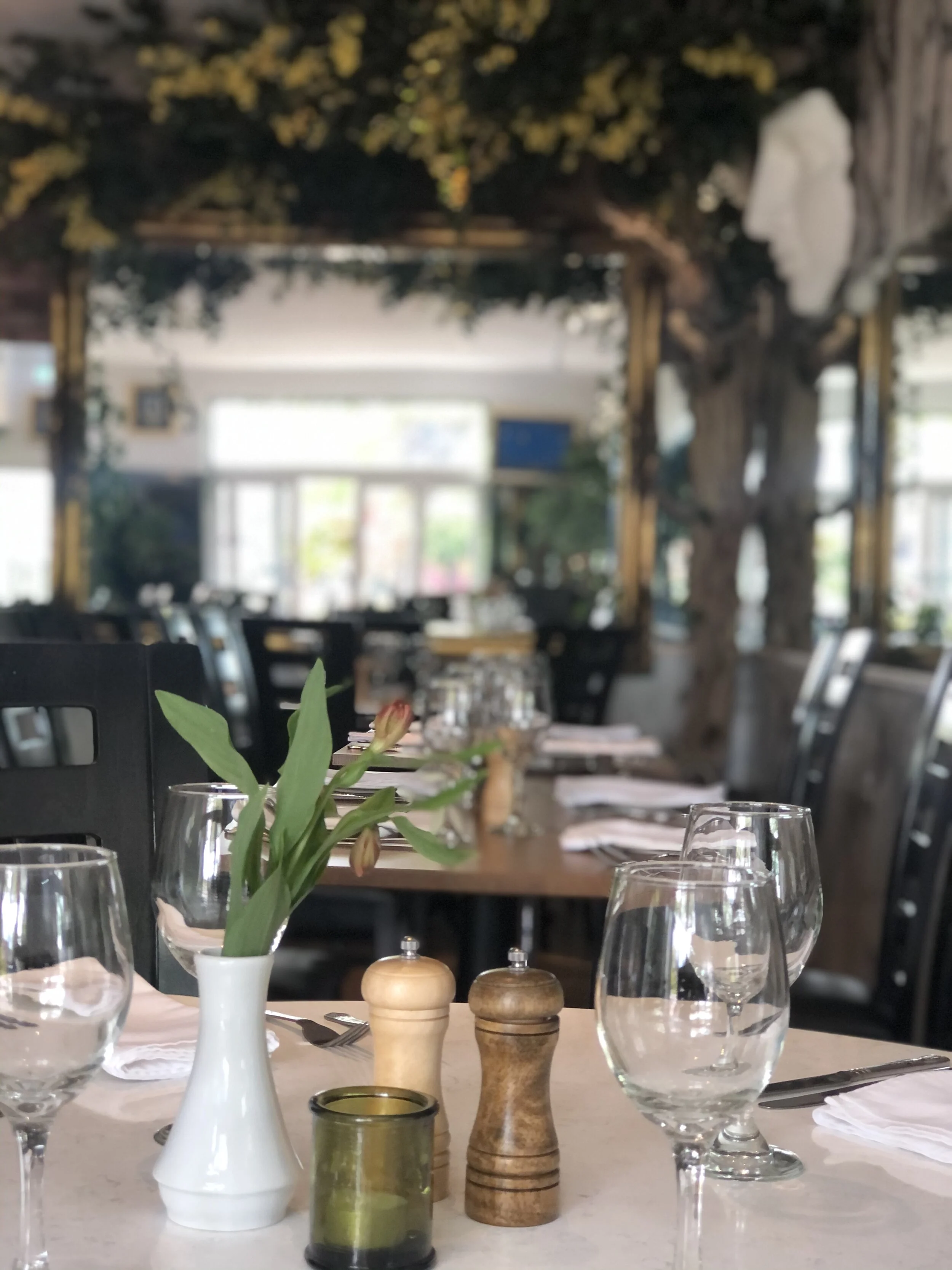 Indoor restaurant dining table decorated with a white vase holding pink flowers, salt and pepper shakers, a green candle holder, wine glasses, and white napkins.
