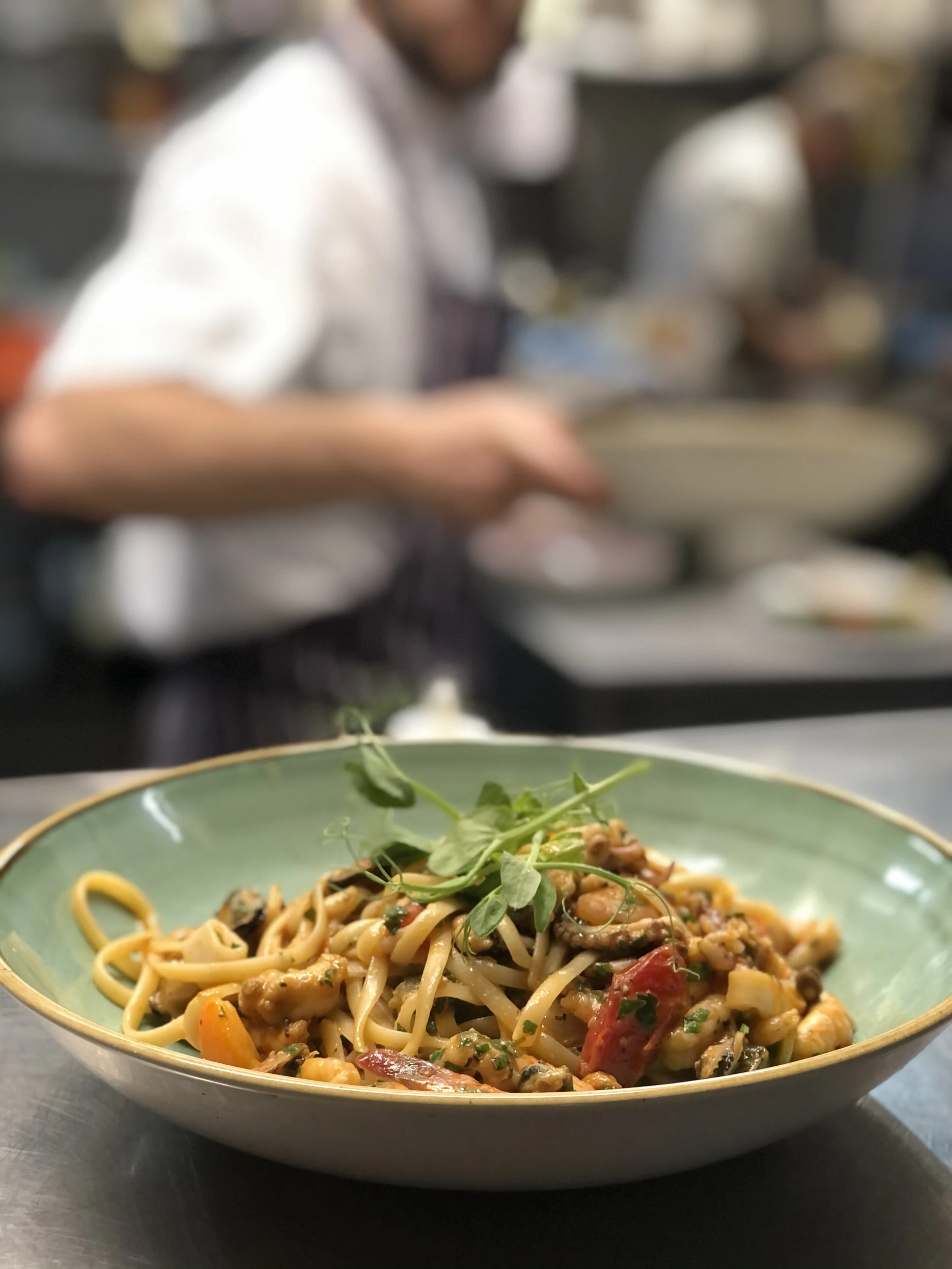 Plate of seafood pasta garnished with microgreens, with a chef preparing food in a commercial kitchen in the background.